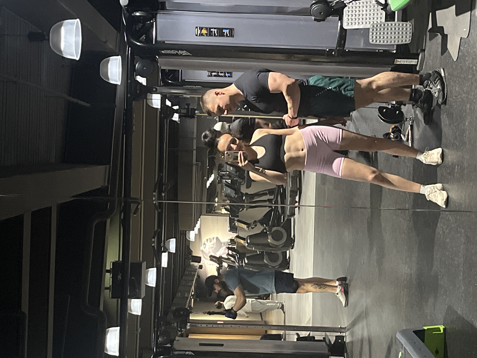 This media file presents an indoor scene within a gym or fitness center, captured with a sideways orientation, making the subjects appear horizontal.
In the foreground, a woman is taking a mirror selfie using a smartphone. She is dressed in a black sports bra and light pink athletic shorts, paired with light-colored sneakers. Her dark hair is styled up. Standing next to her is a man wearing a black t-shirt, dark green athletic shorts, and dark athletic shoes. He has short hair and a noticeable muscular physique.
The background is dominated by a large mirror reflecting the gym interior. Within this reflection, another man is visible, sporting a blue t-shirt, dark shorts, a baseball cap, and athletic shoes. He has a beard. Further back in the reflection, another individual, partially obscured, is seen wearing white attire.
The gym is equipped with various exercise machines, including cable machines and other workout stations, which are visible both directly and in the reflections. The floor is dark, likely rubberized. The space is illuminated by numerous artificial overhead lights, some featuring distinctive semi-circular, bowl-shaped diffusers.
On one of the gym machines reflected in the mirror, there are abstract symbols or icons that appear as "||||" and "T|T|". The overall impression is that of an active fitness environment. Due to the indoor setting and artificial lighting, the specific time of day or external weather conditions are not discernible. The scene is located in Saarbrücken, Germany.