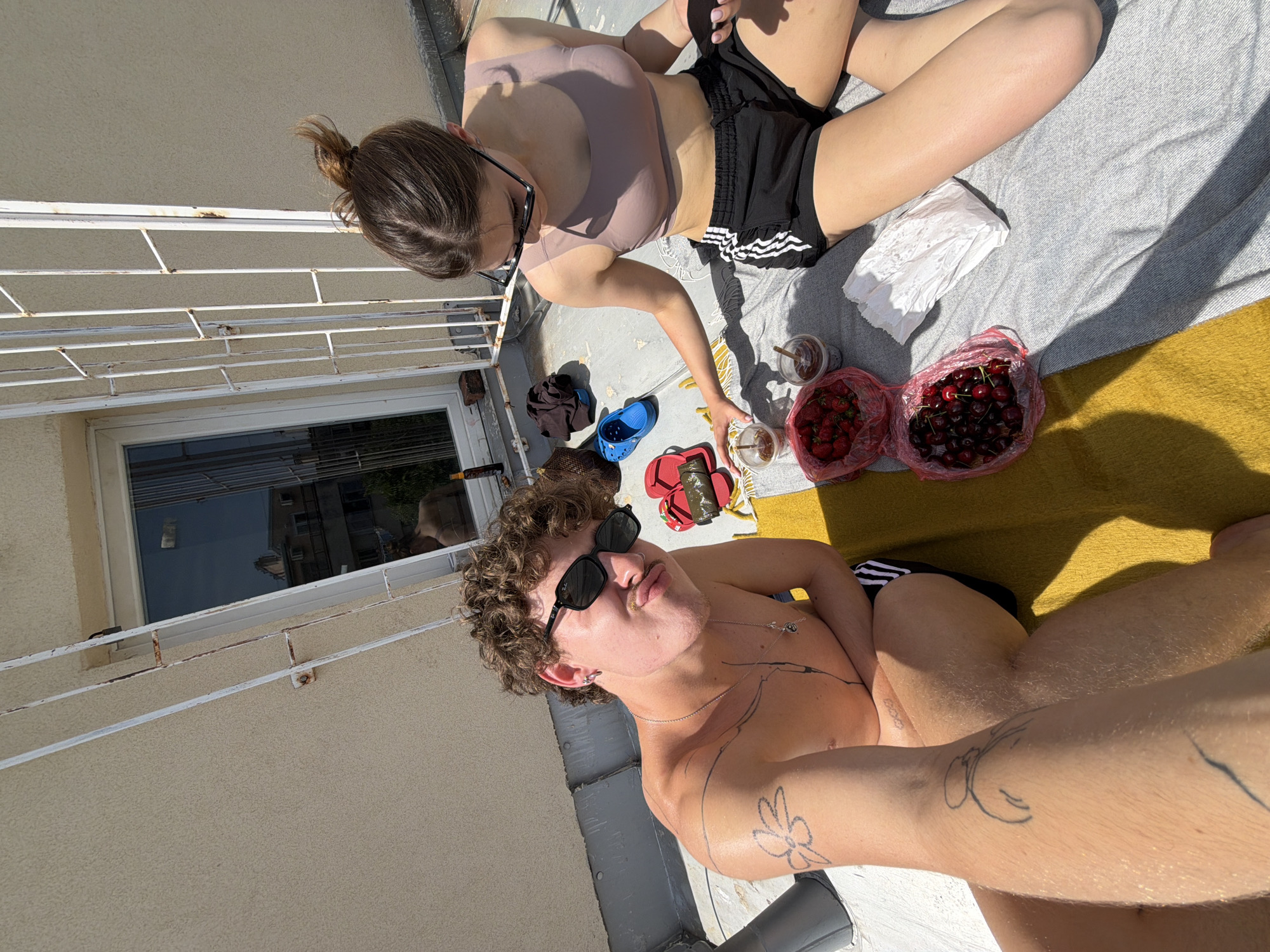 A sunlit, high-angle shot captures two individuals on a rooftop in Vracar Urban Municipality, Serbia. A woman with her hair in a bun, wearing a sports bra and black athletic shorts, is seated on a grey blanket. Beside her are two plastic cups, a bag of strawberries, and a bag of cherries. A man, shirtless and wearing sunglasses, is closer to the camera. He has a flower tattoo on his arm. A white building with a window and a metal railing is visible.
