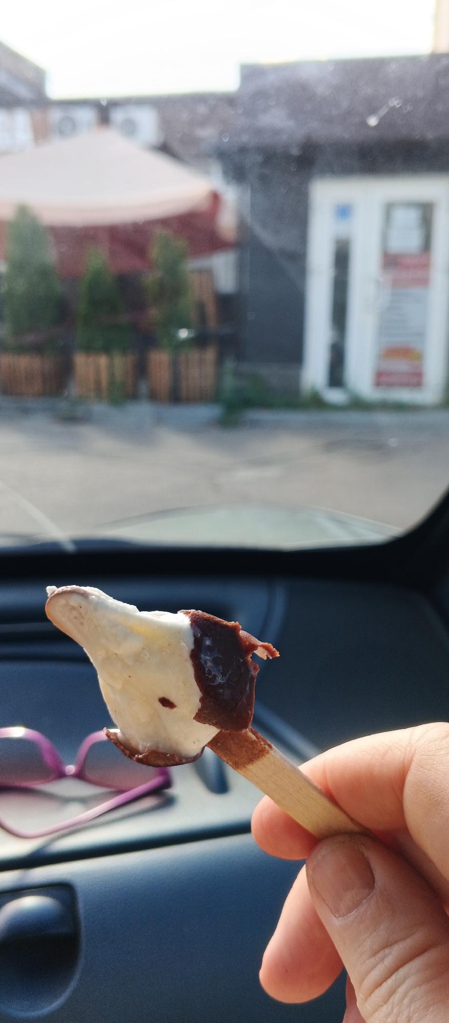 In Smila, Ukraine, a person is holding a partially eaten ice cream bar with a wooden stick in their right hand. The ice cream is white with dark chocolate coating. In the foreground, a pair of pink sunglasses rests on a dark blue surface, possibly a car dashboard. The background shows an outdoor scene with trees in planters, a building with windows, and a light-colored awning. The lighting suggests it is daytime, with sunlight creating shadows and highlights.