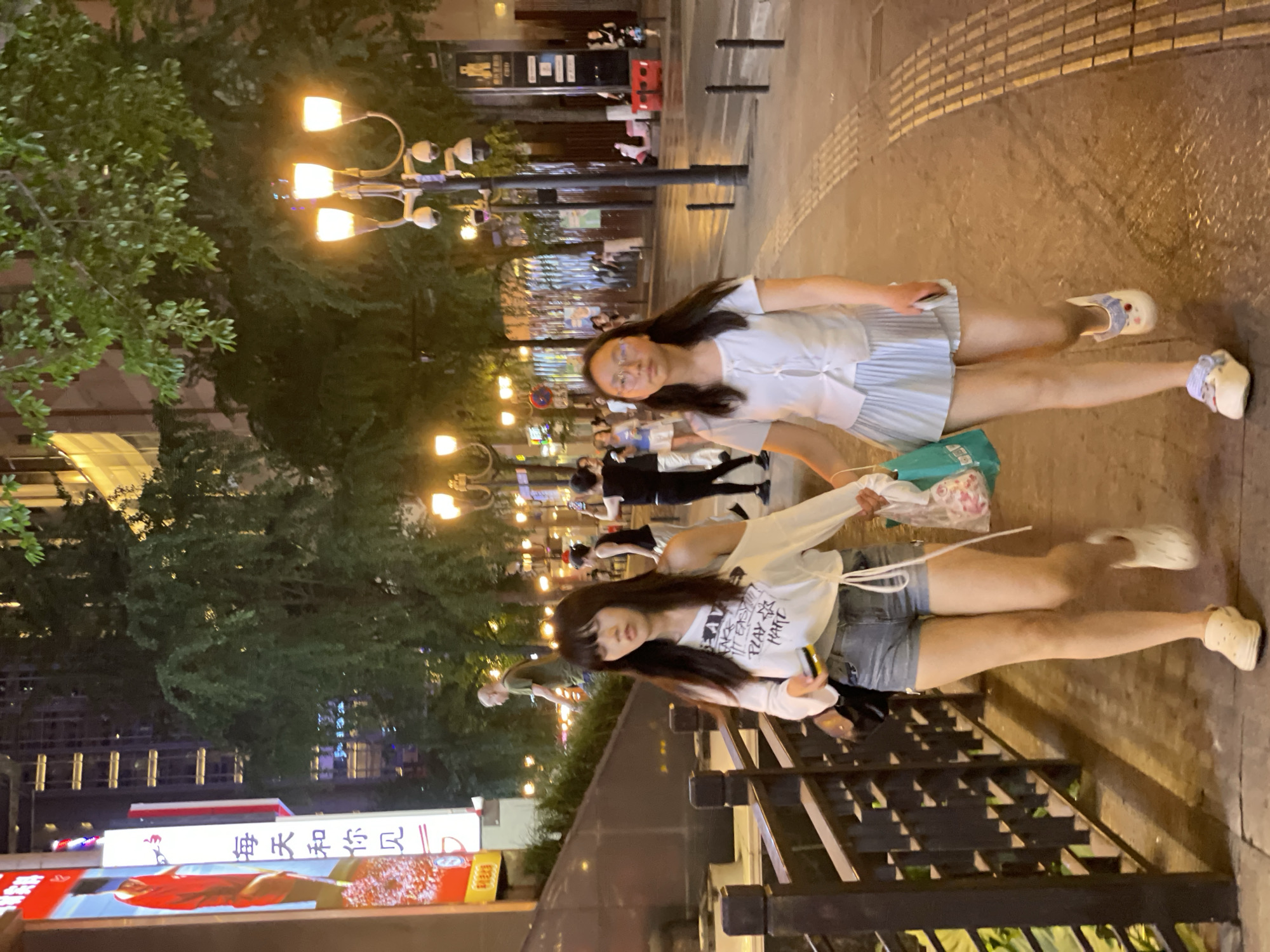 The image depicts a nighttime street scene in Yuzhong District, China, likely a commercial or pedestrian area. Two young women are walking along a paved sidewalk lined with a dark railing on the left and trees with streetlights and buildings in the background. The woman on the left has long dark hair and is wearing an off-the-shoulder white top with black text and graphics, paired with denim shorts and light-colored slip-on shoes. She is holding a phone in her right hand and a plastic bag with pink items in her left. The woman on the right has dark hair and is wearing glasses, a white short-sleeved button-up shirt, and a light blue pleated skirt. She is also wearing light-colored slip-on shoes and carrying a teal bag in her left hand. The sidewalk has textured tactile paving. In the background, there are more streetlights, trees, and the illuminated facades of buildings, including a large vertical sign with Chinese characters and a red graphic on the far left. Other pedestrians and vehicles are visible in the distance.