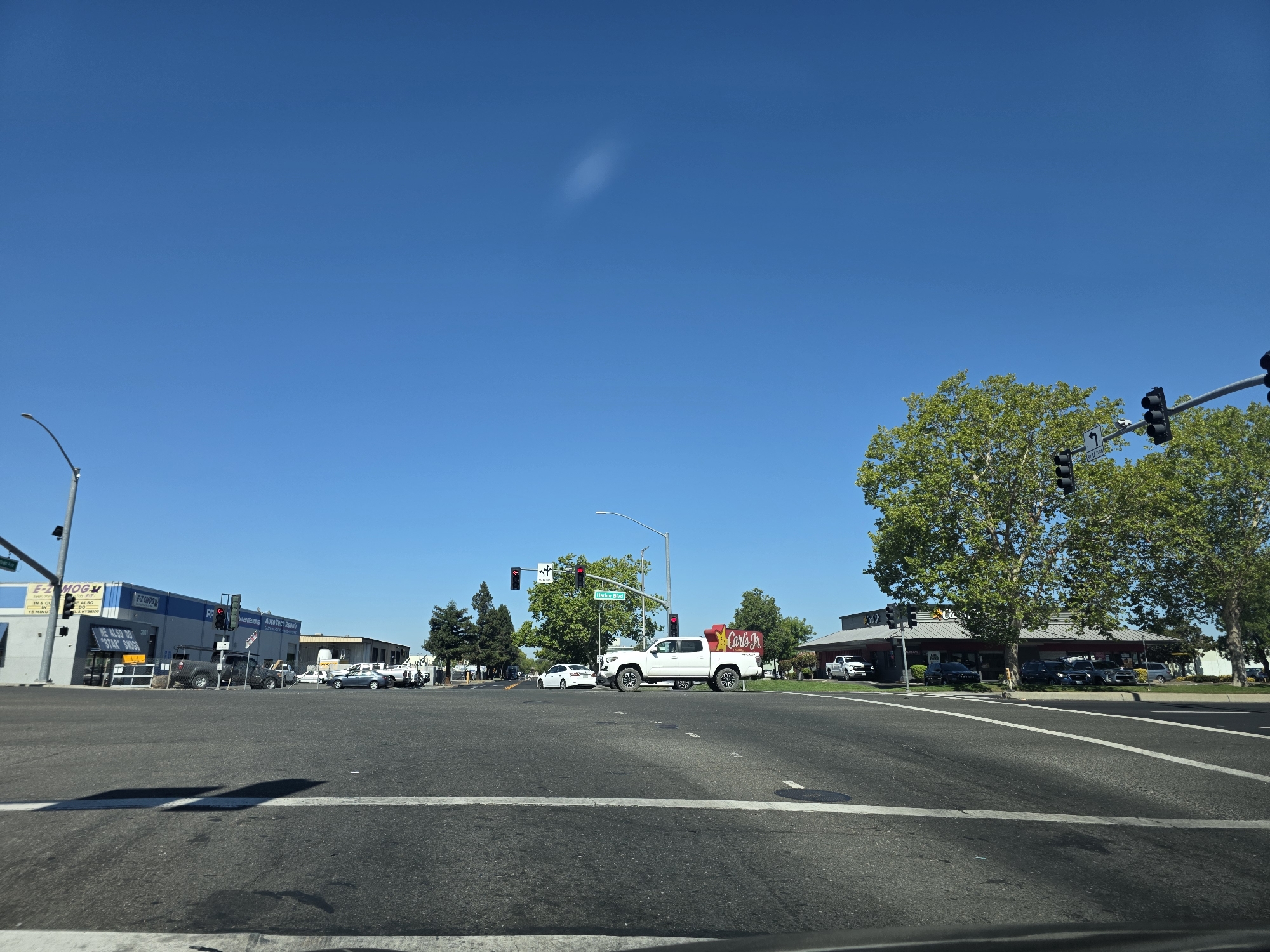 The image depicts a street-level view of an intersection in West Sacramento, California, USA, on a clear, sunny day. The intersection features multiple lanes of asphalt road with white lane markings and a stop line visible in the foreground. Traffic lights are present at the intersection, with a sign indicating "Harbor Blvd".
On the left side of the intersection, a commercial building with blue and gray signage is visible, identifying it as "E-MOG" and "Auto Repair & Service". A dark-colored pickup truck is parked in front of the building. Further down the street, more commercial buildings and trees are seen.
On the right side of the intersection, a white pickup truck with a prominent "Carl's Jr." sign on its bed is stopped at the intersection, facing forward. Behind the pickup truck, a Carl's Jr. restaurant with a red roof is visible, with several cars parked in its lot. Large trees with green foliage are situated in front of the restaurant.
In the distance, more trees and buildings line the street, under a clear blue sky with a faint, wispy cloud. The overall scene is consistent with a suburban commercial area. There are no people clearly visible in the image.