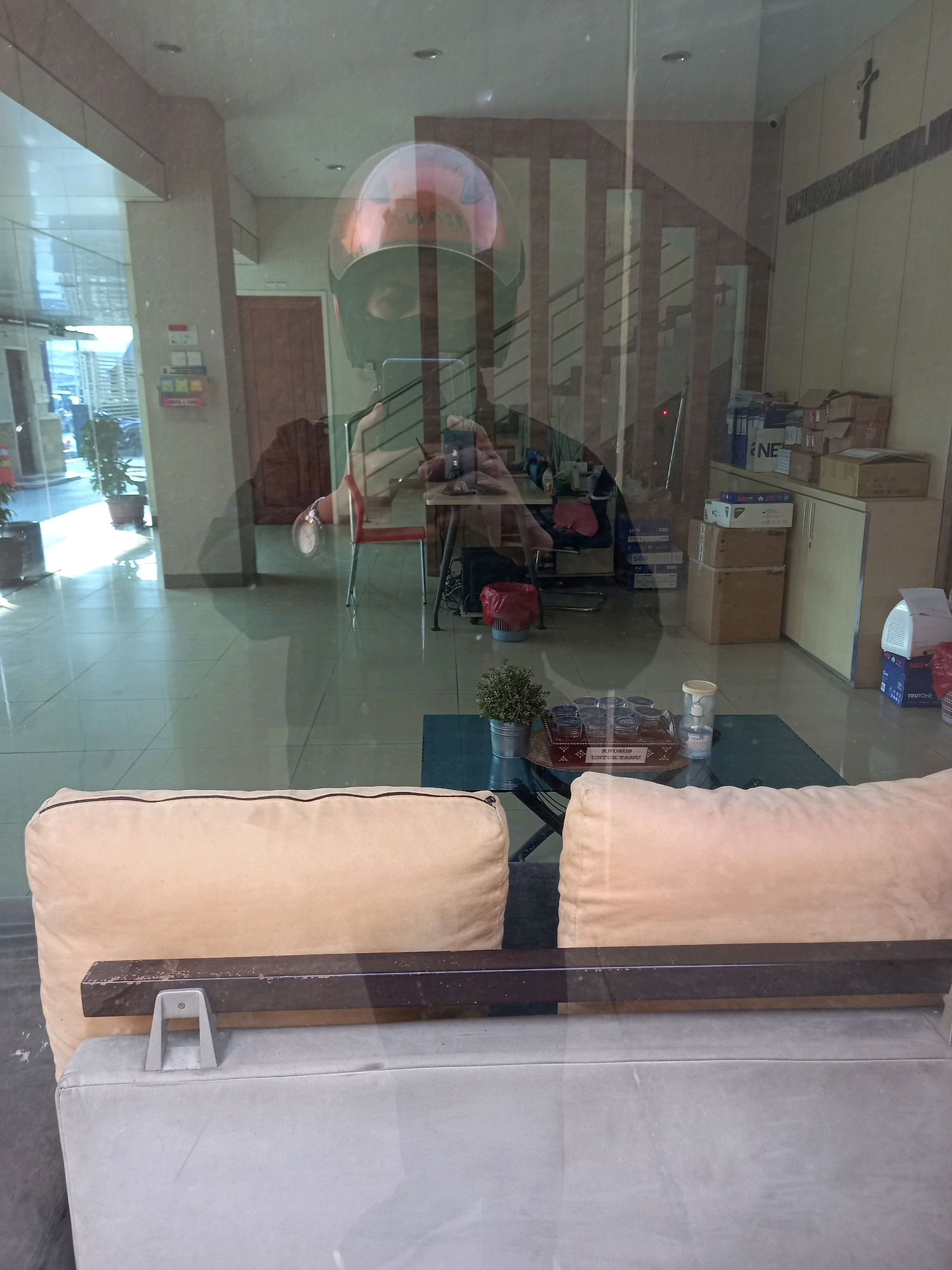 The image depicts a reflection on a glass surface. The reflection shows a person wearing a helmet, holding a smartphone. The environment behind the person includes a reception area with potted plants, a desk with chairs, and office supplies on shelves. In the foreground of the reflection, there are two light-colored sofa cushions. The location is identified as a building in Samarinda, Indonesia, suggested by the presence of Indonesian text visible on a box in the reflection ("KHUSUS UNTUKMU" which translates to "Special for you"). The scene appears to be an indoor office or reception area with a large glass partition or window.