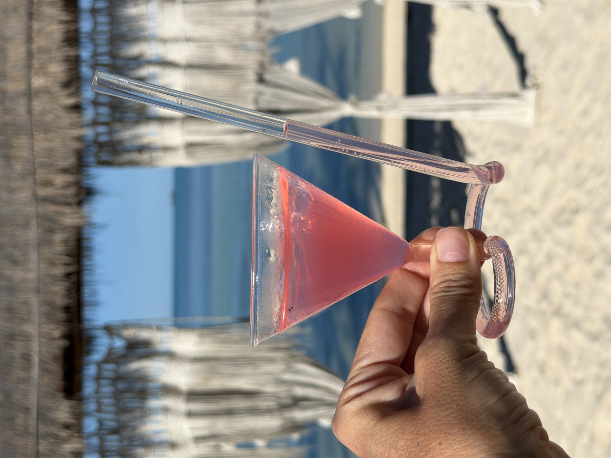 A close-up shot shows a hand holding a pink cocktail in a martini glass with a clear glass straw. The drink is a vibrant pink, with some condensation on the glass. The straw is positioned diagonally across the glass and appears to be made of clear glass, possibly with some subtle pink hue. The background is blurred, but suggests a beach setting with sandy ground, the ocean in the distance, and structures that appear to be thatched roofs and white fabric canopies, commonly found at beach resorts. The lighting indicates it is daytime, with bright sunlight casting shadows. The overall mood is relaxed and suggests a vacation or leisure activity, likely at a tropical location like Baan Thong Krut, Thailand, given the context. There is no visible text in the image.