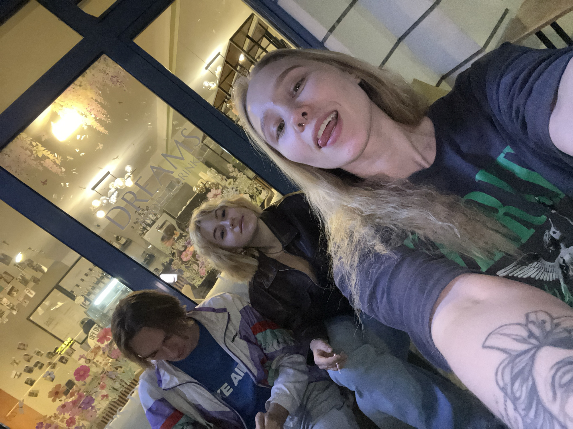 The image shows three young adults in a selfie taken in front of the "Dreams Drink" bar in Budva, Montenegro. A young woman with long blonde hair and a nose ring takes the selfie, sticking her tongue out towards the camera. Her arm is visible in the foreground with a tattoo. Next to her is another young woman with blonde hair in a dark leather jacket holding a cigarette. To the left is a young man with brown hair wearing a blue t-shirt and a white jacket with blue and purple accents. The background shows the front window of the "Dreams Drink" bar, which is adorned with decorations and photographs.
