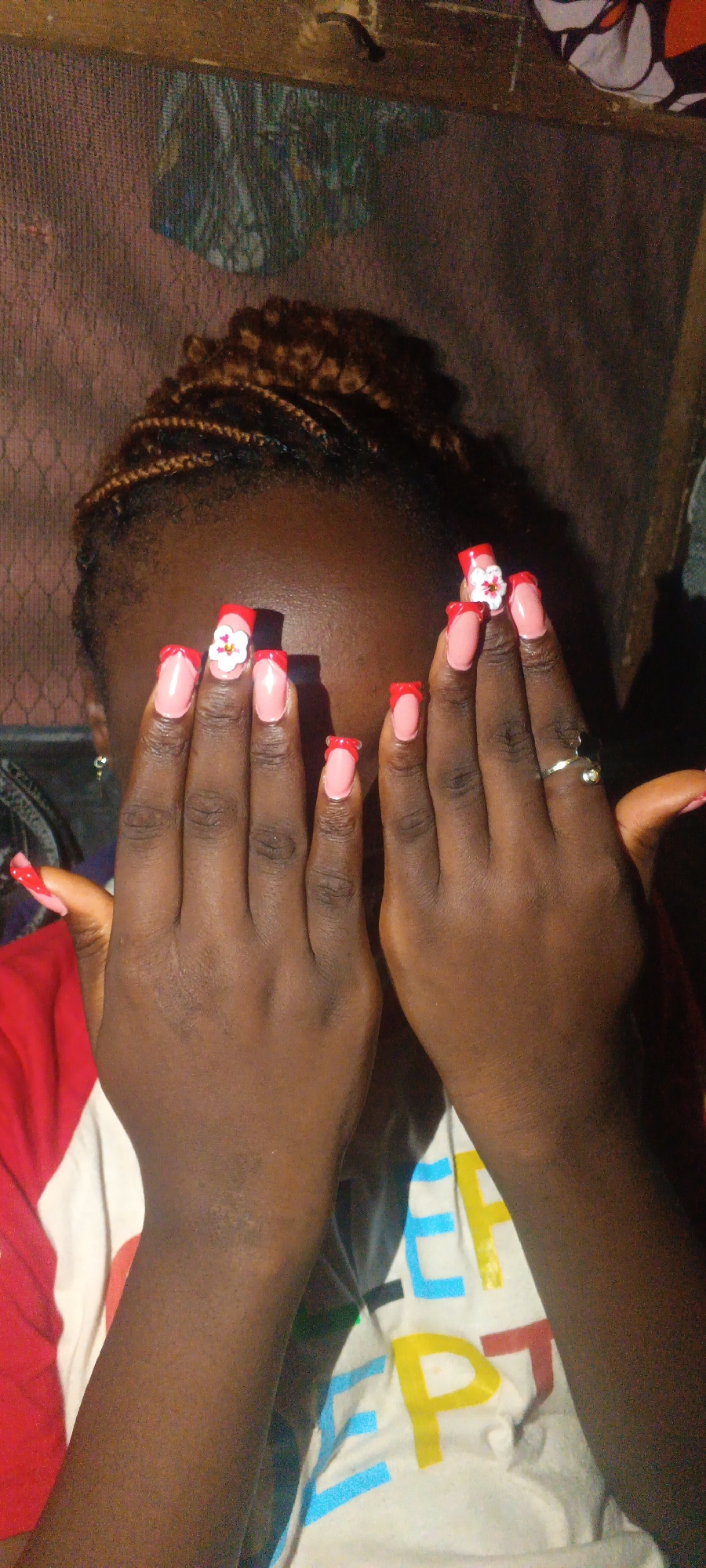 A person is photographed indoors, holding up both hands to obscure their face. The person appears to be dark-skinned, with brown braided hair. The nails are long and painted pink with a red French tip, with a flower embellishment on one nail. The person is wearing a shirt with a red sleeve and a white front with colorful lettering. A gold ring is visible on the right hand. The background is indistinct, with a patterned wall visible behind the person's head. Based on the low lighting and background elements, it is not possible to confirm the location in Onitsha, Nigeria.