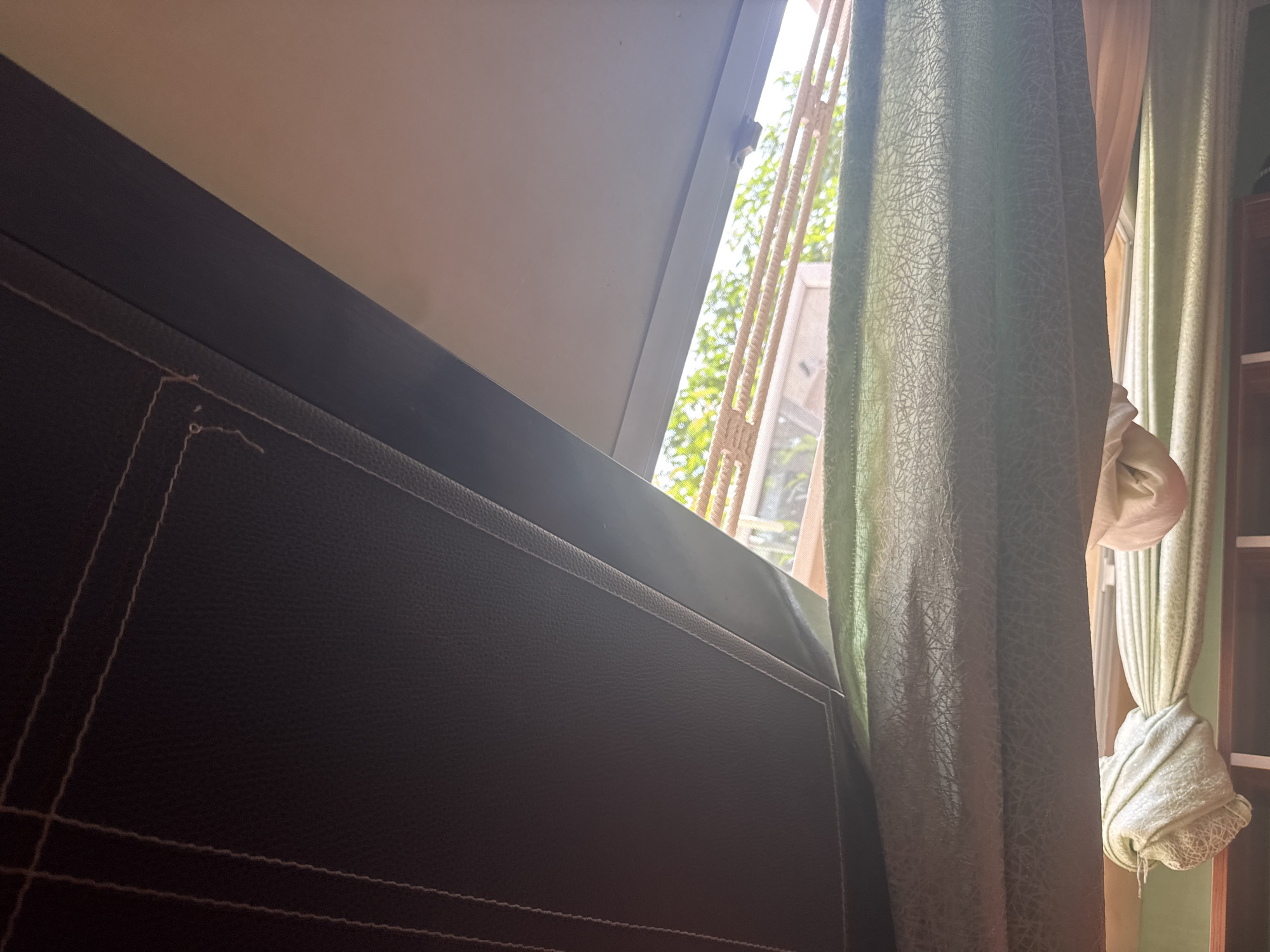 The image captures a close-up view of a dark brown, textured surface, likely upholstery, with visible stitching forming geometric patterns. To the right of this surface, a window is partially obscured by light green and cream-colored curtains. The curtains are draped and tied back in a way that reveals a glimpse of the outdoors through a barred window. Sunlight streams through the window, creating bright highlights and casting a soft glow on the scene. The foliage of trees is visible outside, suggesting a daytime setting with pleasant weather. The overall impression is one of a domestic interior, possibly a bedroom or living area, looking out onto a green, natural environment. The lighting suggests it is daytime, perhaps late morning or early afternoon. There is no visible text in the image. The mood is calm and peaceful, with the natural light creating a warm and inviting atmosphere. The location context of Narayi, Nigeria, implies a tropical or subtropical climate where such greenery and sunlight are common. The barred window could indicate security measures typical in some regions.