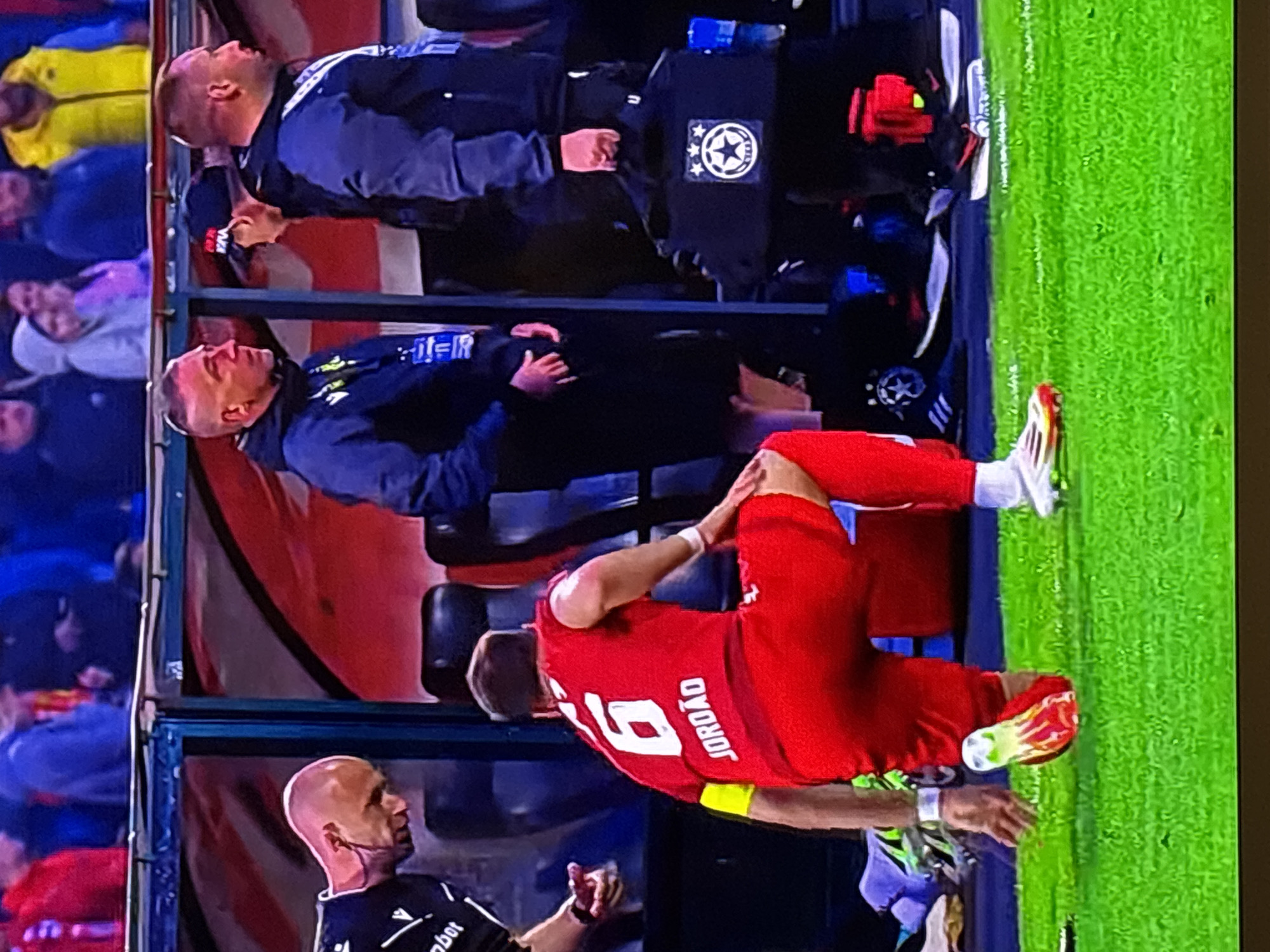 The image captures a nighttime scene at a football (soccer) stadium, focusing on the technical area and a player on the field during what appears to be a professional match. The artificial lighting indicates an evening or night game.
In the foreground, a player wearing a red jersey with the number "19" and the name "JORDAO" on the back is kneeling on the green grass field, resting on his hands with one knee bent. He is wearing distinctive red, white, and yellow/green football boots, suggesting a moment of pause or slight discomfort.
To the player's left, a bald official is standing, wearing a dark shirt with the text "bit" or "robot" partially visible, and a headset with a microphone. He is looking towards the player.
Behind the sideline, two men, likely coaches or team staff, are seated on a red bench. Both have short hair or are bald and are dressed in dark jackets. Prominently displayed on a piece of equipment behind the upper man on the bench is a clear UEFA Champions League logo, featuring a white star with three smaller stars inside. This detail confirms the context of a high-profile European club football competition.
The background shows the blurry outlines of spectators in the stadium stands, illuminated by the stadium lights. The scene depicts a typical moment during a professional football match, possibly an injury assessment or a brief break in play. The presence of the UEFA Champions League logo strongly suggests the event is part of this competition, potentially taking place in Sofia, Bulgaria, as per the provided context.