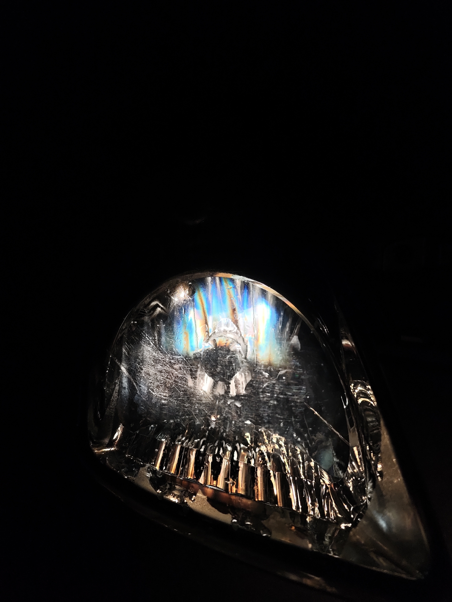 The image depicts a close-up view of a car headlight. The headlight appears to be slightly scratched and has a rainbow-like diffraction pattern across its upper surface. The lower part of the headlight has a series of vertical, reflective elements. The background is completely black. There is no information to suggest this image was taken in Kozloduy, Bulgaria. There are no people or interactions visible.