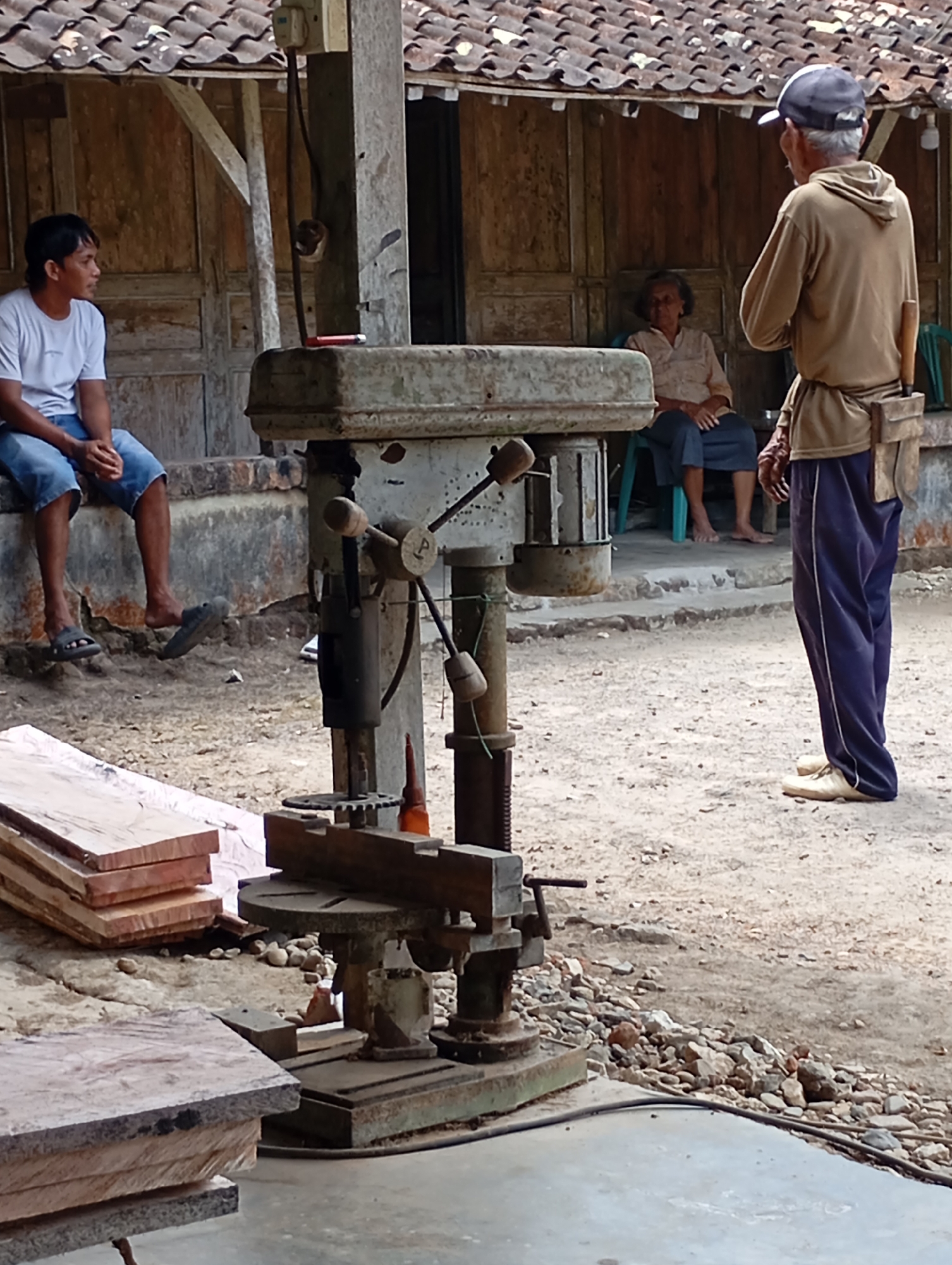 In Ketepung, Indonesia, an outdoor scene unfolds with a focus on a vintage drill press. A young man in a white t-shirt and blue shorts sits on a low concrete ledge, facing left, his hands clasped. Beside him, to the right, a weathered drill press stands as the central object, its metallic components showing signs of age. Two older individuals are visible in the background. An elderly woman in a light-colored patterned shirt and dark pants sits on a green chair, facing towards the drill press. To her right, a man wearing a tan hooded sweatshirt, dark blue pants with white stripes, and a blue baseball cap stands facing the woman and the drill press. He has a tool pouch attached to his belt. The environment suggests a rustic setting with a wooden building with a tiled roof in the background. Stacked lumber is present in the foreground to the left, and scattered rocks litter the ground around the drill press, indicating a possible workshop or outdoor work area.