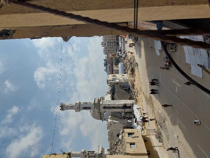 The image depicts a street scene in Khan Yunis, Palestinian Territory, characterized by widespread structural damage under a partially cloudy blue sky. The vantage point is from a damaged building, with part of a dark, wrought-iron structure visible in the immediate right foreground.
In the mid-ground, a wide, unpaved or dust-covered street extends into the distance. Several individuals and groups are present on the street. A group of approximately six to seven people in dark clothing walks collectively towards the viewer's right. Further down the street, a solitary figure in a reddish shirt walks away from the viewer. Another person, carrying a white, rectangular object, walks on the left side of the street towards the background. Near the bottom-center, a small white object, possibly a container, rests on the street. On the right side of the street, a makeshift stall or cart with various small items is visible. White tarps or coverings are spread on the ground near the bottom right. Faint arrow markings are present on the road surface.
The background is dominated by a large, white mosque featuring a dome and two minarets. The mosque exhibits extensive damage, with significant portions of its structure, including parts of its walls and roof, collapsed or missing, revealing internal debris. Surrounding the mosque and lining the street are numerous multi-story buildings, many of which are heavily damaged, showing exposed interiors, shattered windows, and collapsed sections. Large piles of rubble and debris are scattered along the sides of the street and around the damaged structures. A thin, dark cable or wire traverses horizontally across the upper portion of the scene.