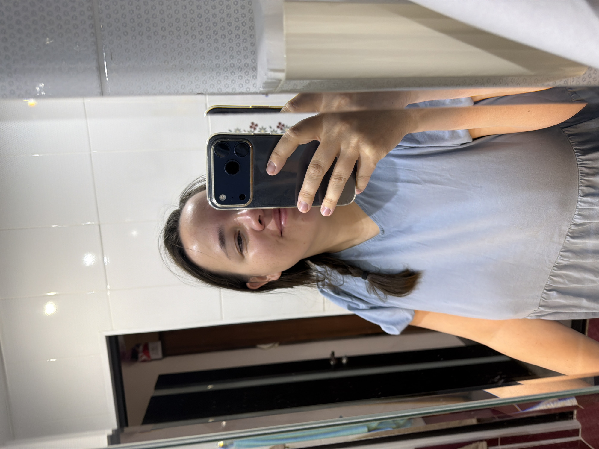 The image, rotated 90 degrees clockwise, depicts a mirror selfie taken by a young woman indoors, likely within a room featuring white tiled walls, in Chaikovsky, Russia. The woman, who has long dark hair styled with a visible braid on her left side, is wearing a light blue short-sleeved top or dress. She holds a dark-colored smartphone horizontally with her right hand, using it to capture her reflection. The phone is encased in a protective cover that features a metallic, possibly gold-toned, rim around its prominent multi-lens camera system.
The woman's face, with a neutral expression, is turned towards the phone as she takes the picture. Her complexion appears natural, without noticeable heavy makeup.
The background reflected in the mirror primarily consists of white square tiles with visible grout lines, suggesting a bathroom or similar area. Above the tiled section, a lighter area with a subtle, repetitive circular or floral pattern is reflected, possibly from wallpaper or a ceiling. Below the woman's reflected torso, a dark, horizontally oriented surface, likely a countertop or part of a vanity, is visible. Further in the reflection, a darker interior space with indistinct furniture or shelving can be faintly perceived. Bright light reflections on the tiles indicate artificial lighting. There is no visible text in the image, and the specific time of day or outdoor weather conditions cannot be determined from the indoor setting.