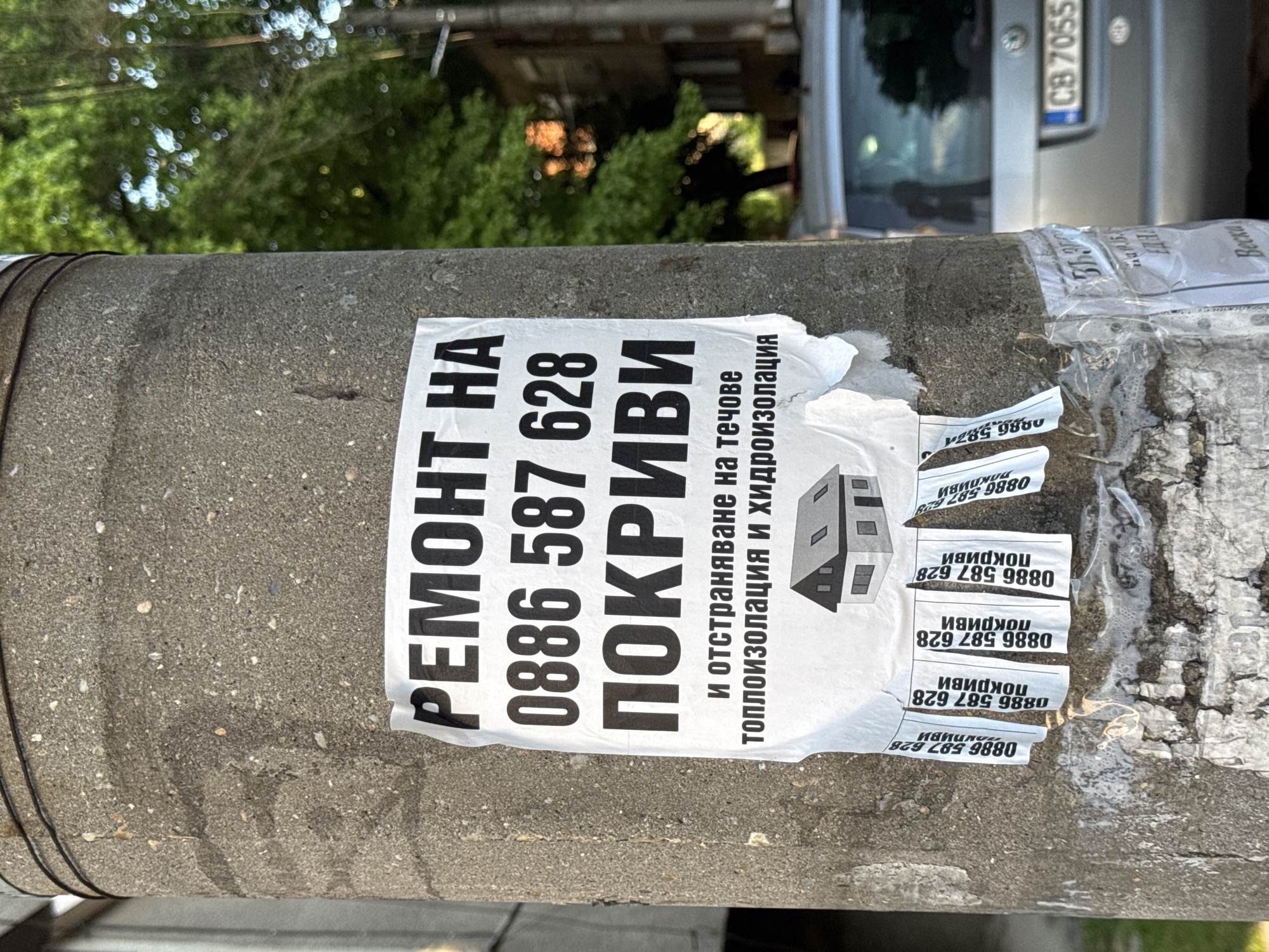 A close-up shot shows a concrete pole covered in advertisements. A primary advertisement, in Bulgarian, states "REPAIR OF ROOFS" followed by the phone number "0886 587 628", also mentioning "waterproofing and thermal insulation". The pole has multiple torn remnants of the same advertisement stacked on top of each other. A glimpse of a vehicle with a license plate "CB 7055" can be seen in the background. The surrounding environment is blurred, showing green foliage.