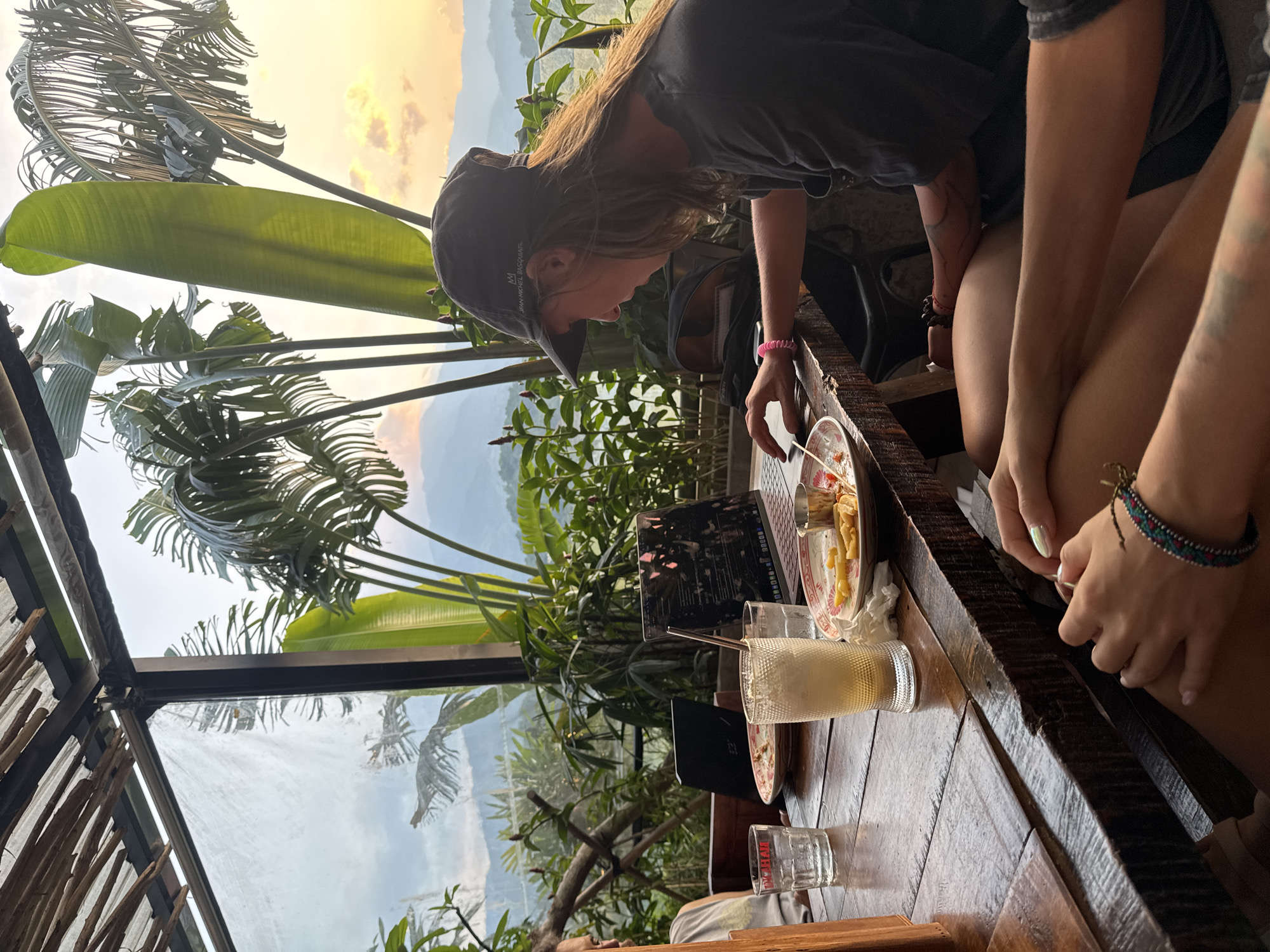 A daytime shot shows two people seated at a wooden table on an elevated platform, possibly in a restaurant or cafe, with a view of the Hà Giang mountain range and sky visible in the background. The location appears to be in Hà Giang 1 Ward, Vietnam, indicated by the distinct geography and likely vegetation.
The person on the left is a young adult, possibly female, wearing a black baseball cap and a dark t-shirt. She is leaning forward, interacting with a laptop on the table, and has a pink bracelet on her left wrist. The person to her right has tattooed arms and is wearing a dark t-shirt and shorts. They are holding a small item in their hands.
On the table are plates with leftover food, a beverage in a tall glass, several empty glasses, a laptop, and other small items. Lush greenery is visible outside the enclosure, further suggesting a tropical or subtropical location. The setting is lit naturally, with the sky suggesting either dawn or dusk. The elevated structure appears to be part of a greenhouse-like construction, with a glass or plastic roof.