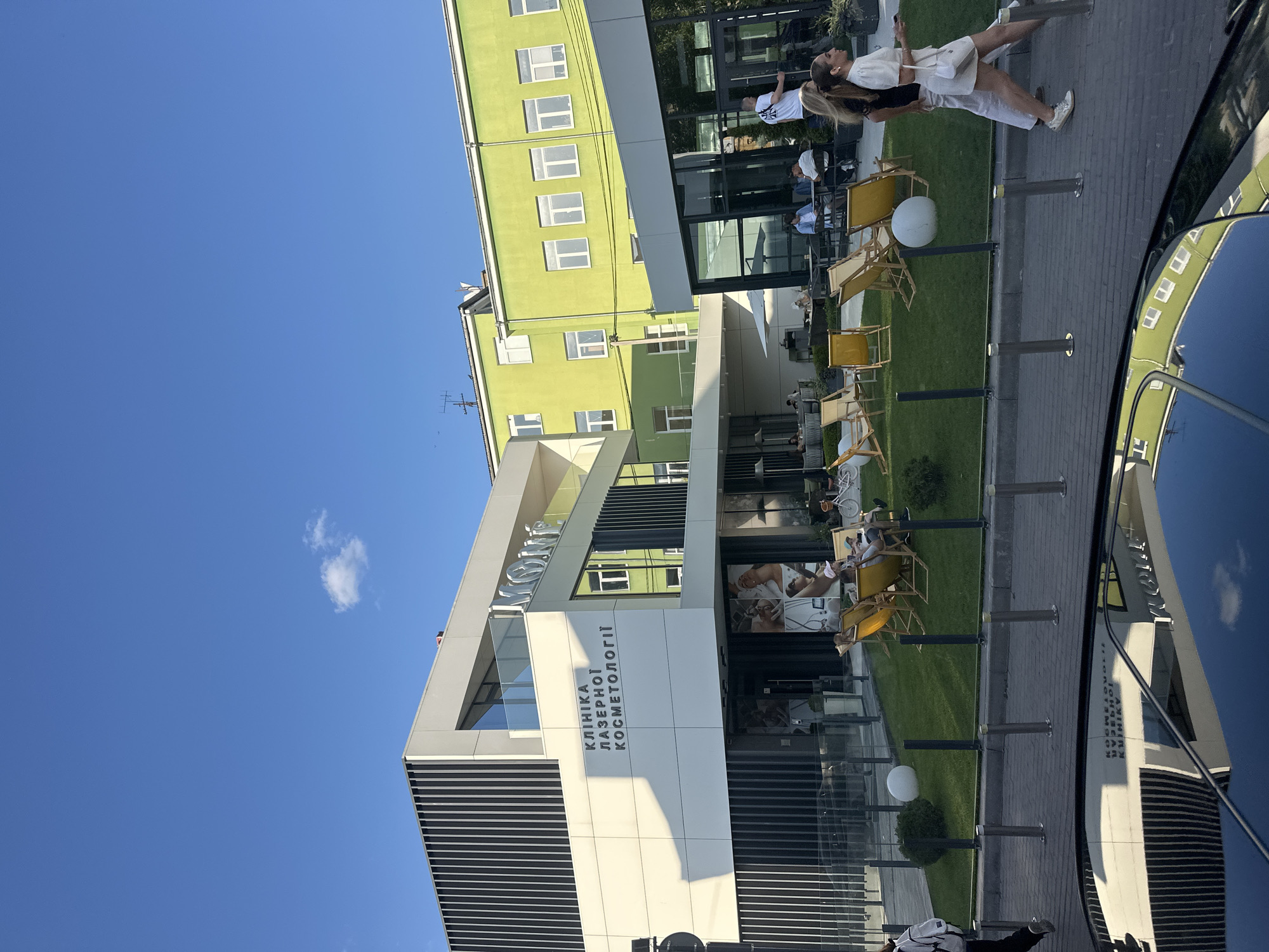 The image depicts the exterior of a modern building complex in Ternopil, Ukraine, identified by signage as "КЛІНІКА ЛАЗЕРНОЇ КОСМЕТОЛОГІЇ" (Clinic of Laser Cosmetology) and a large white "MON" logo. The building features clean lines, white and grey facades with vertical black slats, and glass windows. Adjacent to it is a taller, bright green building with multiple white-framed windows.
In front of the clinic, a well-maintained lawn area is set up with yellow lounge chairs, white spherical decorations, and some landscaping. Several individuals are seated in the lounge chairs, some appearing to relax or engage in conversation. One person is standing and walking away from the camera.
In the foreground, the reflection of the clinic building and the green building is visible on the dark, glossy surface of what appears to be a car's roof or hood. The sky is clear blue with a single small white cloud. The overall scene suggests a bright, sunny day in an urban setting, likely during daytime hours.