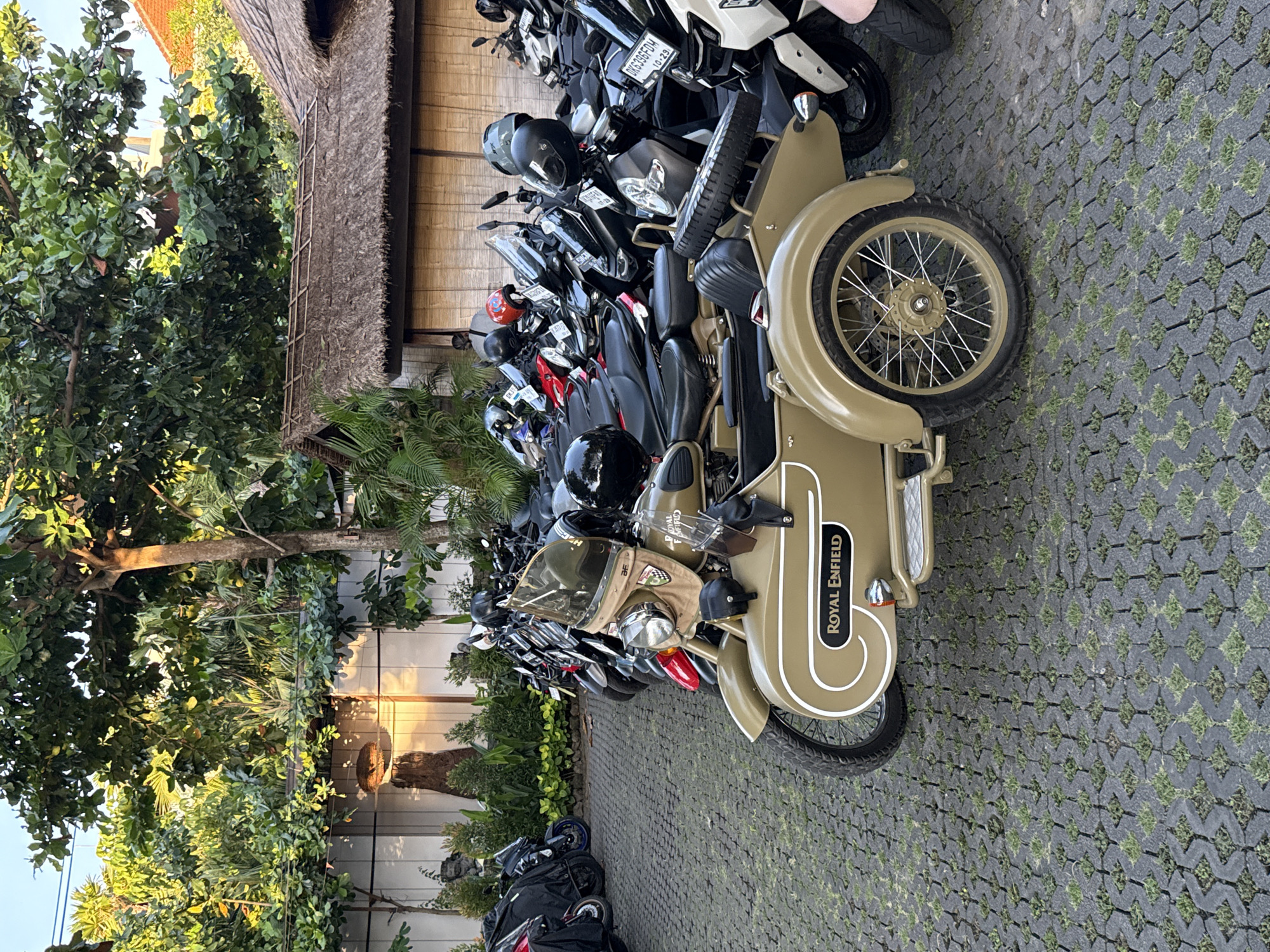 The image depicts a row of parked motorcycles and scooters in Kuta Selatan, Indonesia. A Royal Enfield motorcycle with a sidecar is prominently featured in the foreground. The ground is paved with interlocking concrete blocks with grass growing in the gaps. Lush tropical foliage and trees are visible in the background, along with a building with a thatched roof. The lighting suggests it is daytime.