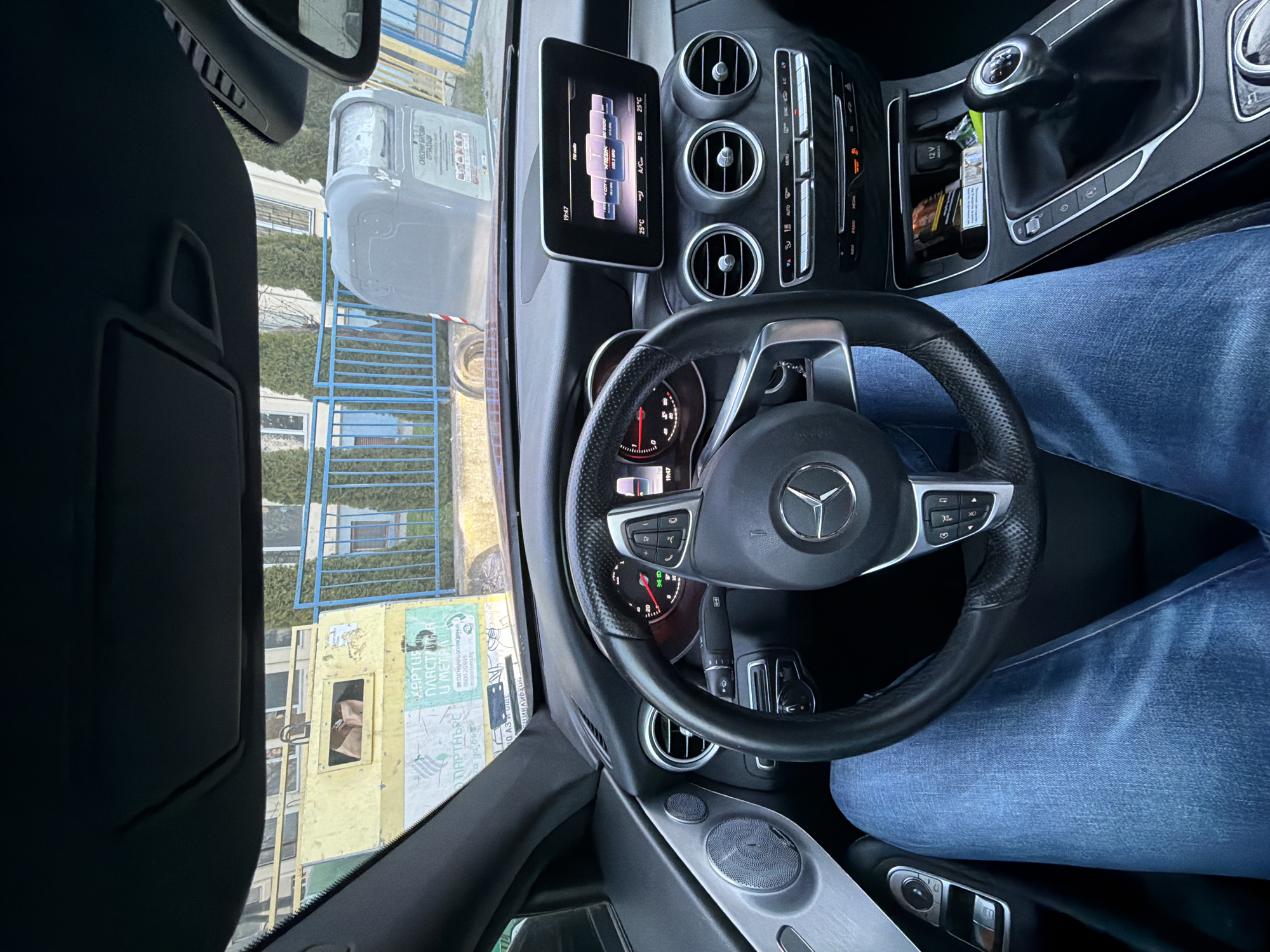 The image depicts the interior of a Mercedes-Benz car, focusing on the driver's perspective. The steering wheel, dashboard with a digital display showing the time and temperature (19:47, 25°C), and center console are visible. The gear stick and surrounding area are also in view, with a pack of cigarettes visible inside the 12v socket. The driver is wearing blue jeans.
Through the windshield, we can see a glimpse of the outside environment. There's a blue metal fence and what appears to be a yellow trash container. The container has some text in Bulgarian, including "ХАРТИЯ ПЛАСТМАСА И МЕТАЛ" (Paper, Plastic and Metal) and "СМЕСЕНИ БИТОВИ ОТПАДЪЦИ" (Mixed Household Waste), as well as a phone number. Based on the Bulgarian text and the image, it is possible that the location is in Varna, Bulgaria, although there's no definitive indicator confirming that.