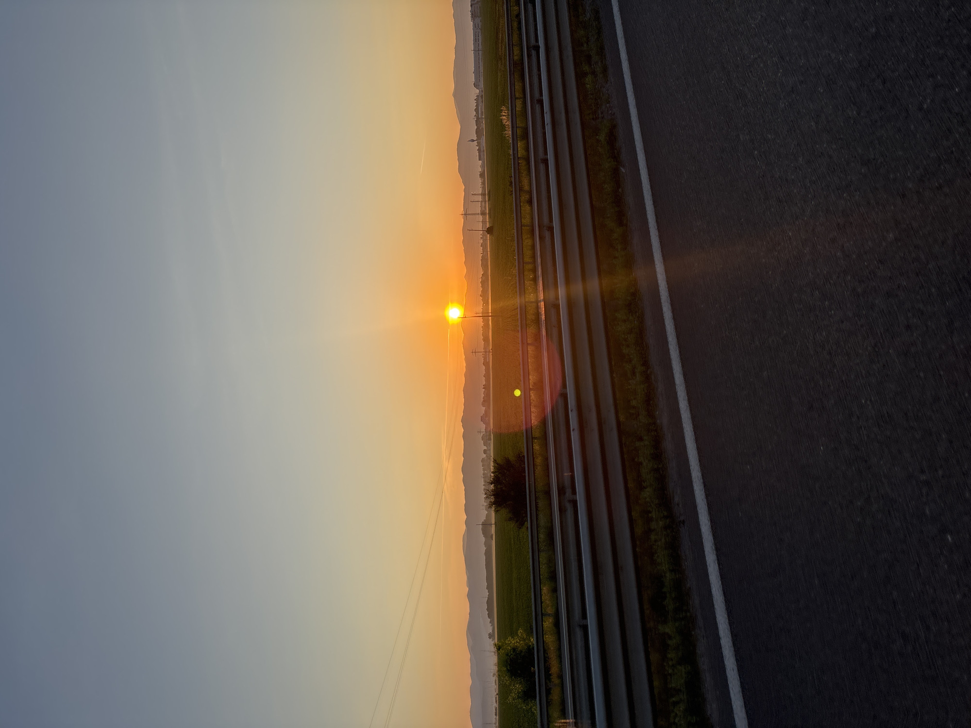 The image depicts a highway scene at sunrise near Sofia, Bulgaria. The sun is low on the horizon, casting a warm orange glow across the sky and landscape. In the foreground, a dark asphalt road is visible, with a white line marking the lane. Beyond the road, a metal guardrail separates the highway from a green field. In the distance, rolling hills or mountains form a hazy silhouette against the bright sky. Power lines and utility poles are scattered across the landscape, indicating the presence of infrastructure. The overall atmosphere is serene and tranquil, characteristic of an early morning journey.