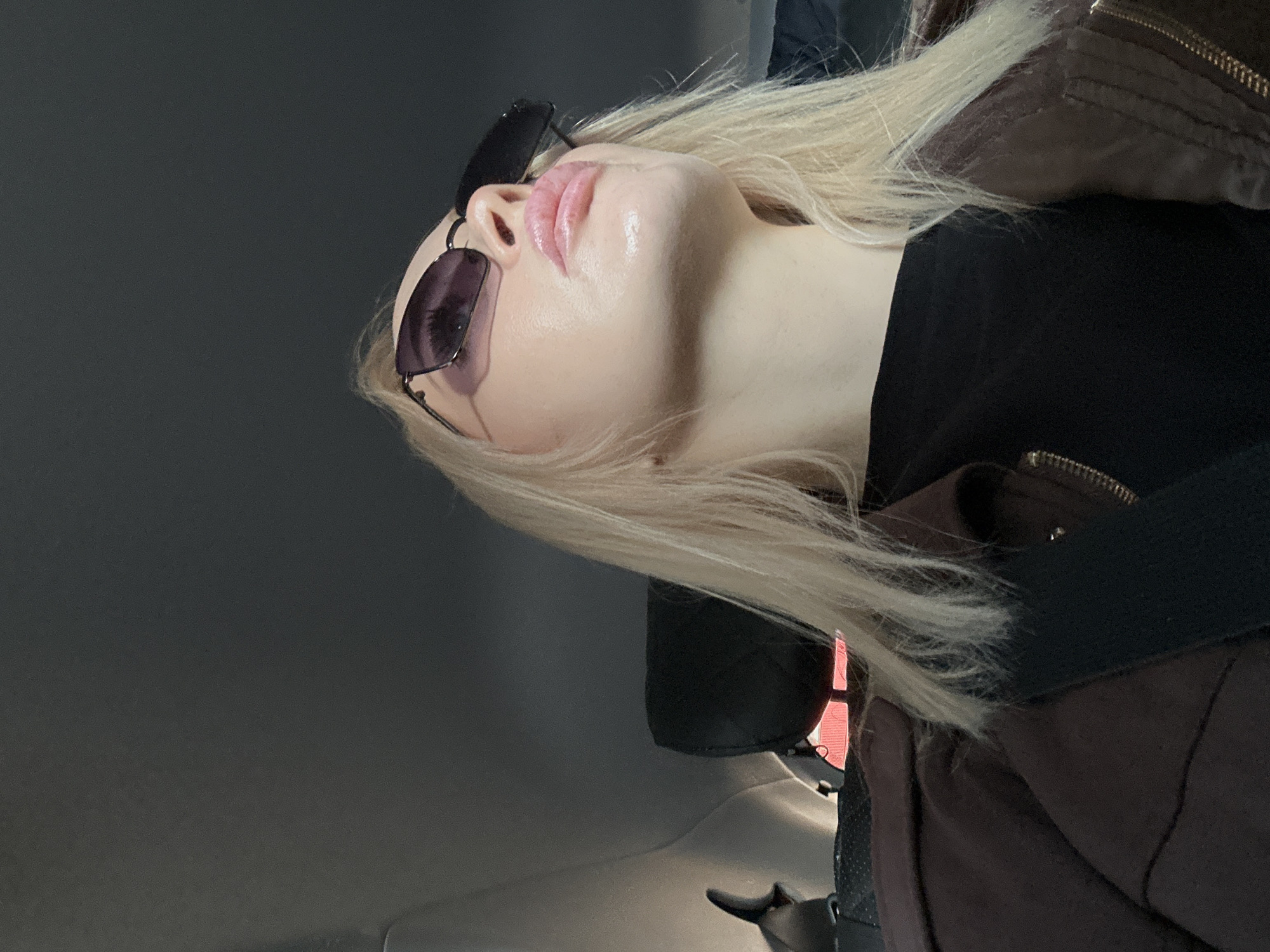 A fair-skinned individual with blonde hair is wearing dark sunglasses, a black top, and a brown jacket, with a black seatbelt crossing their body. The image appears to be taken inside a vehicle, possibly a car, with a gray interior roof. The location of Megion, Russia, is not discernible from the image.