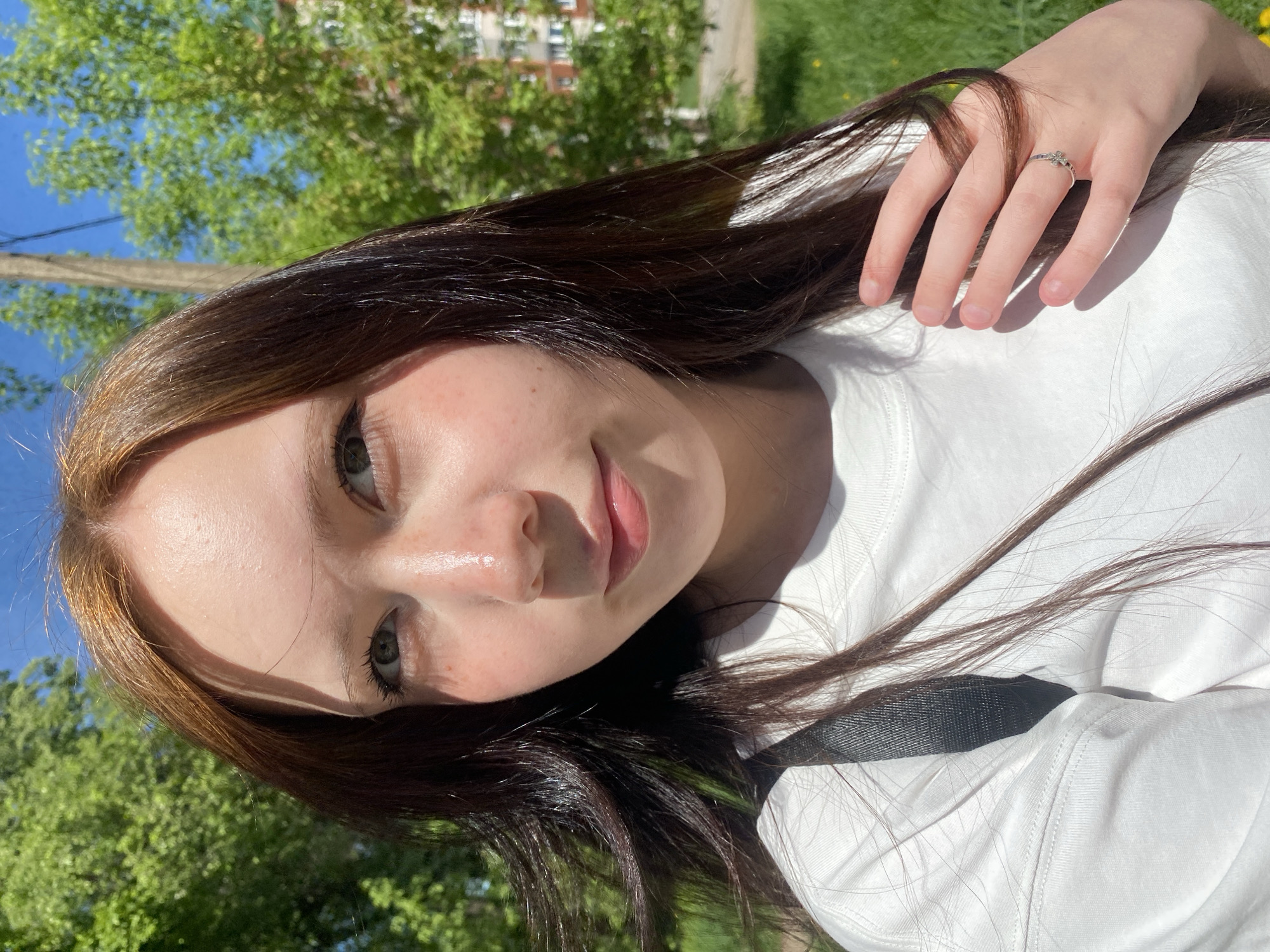 A person is facing the camera, with her head turned slightly to the left. She has brown hair and light skin. She is wearing a white shirt. Her right hand is raised towards her face. She is outdoors, with green trees and a building visible in the background. The location is Unknown, Unknown.
