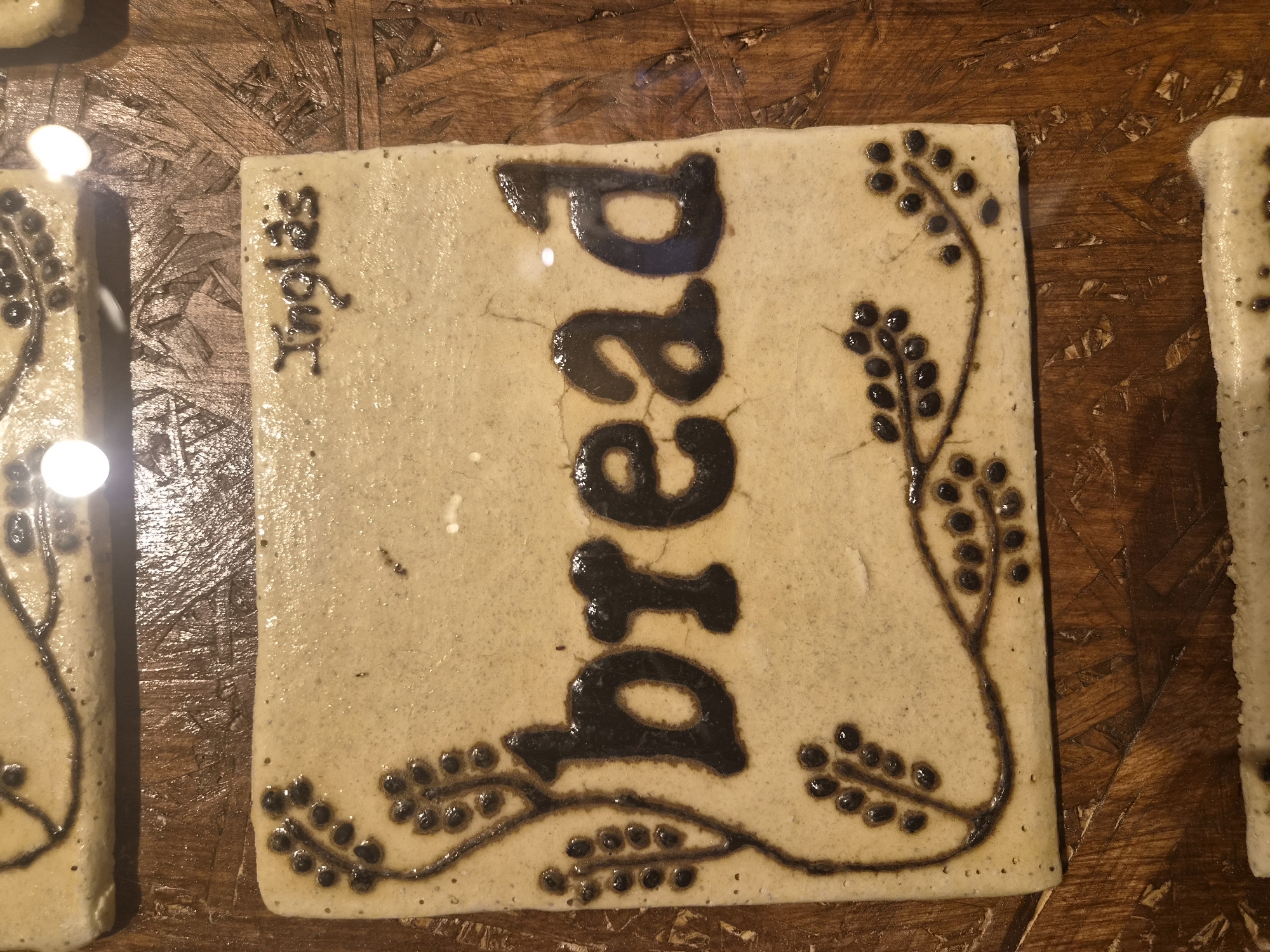 The image shows a close-up of two ceramic tiles on a wooden surface. The top tile is partially visible and has a decorative pattern. The bottom tile is rectangular and features the word "bread" in large, dark letters in the center. Above the word "bread," the word "Inglês" is inscribed in smaller, darker letters. The tile is adorned with a dark brown decorative design that resembles branches with berries, flanking the word "bread" on the left and extending across the bottom. The background is a textured wooden surface, likely a table or countertop. The lighting appears to be artificial, casting a slight glare on the glossy surface of the tiles. The context suggests a shop or display of artisanal food-related ceramic items, possibly in Seia, Portugal, given the presence of the Portuguese word "Inglês" (English). There are no people or actions depicted in the image.