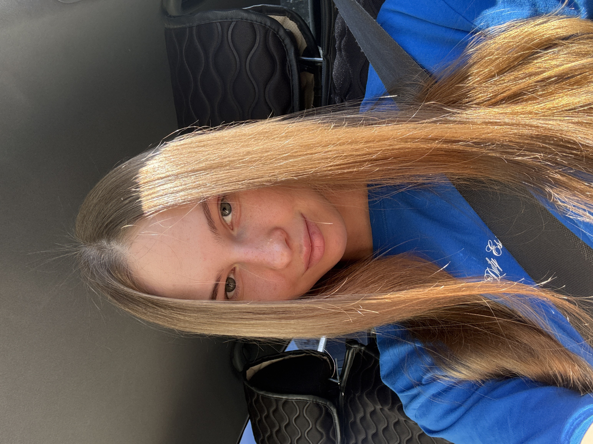 This is a close-up, portrait-style photo of a young woman taken from a low angle. She has long, blonde hair that is slightly windblown, and her head is tilted to the side. She is wearing a bright blue t-shirt with white script text on it, partially obscured by her hair. A dark gray seatbelt is visible across her chest. The background is blurred, but appears to be the interior of a car, with textured black seat covers visible. The lighting suggests it is daytime, possibly outdoors, with sunlight catching the woman's hair and face, creating a warm glow. Her expression is relaxed and natural. The location context is Perm, Russia.