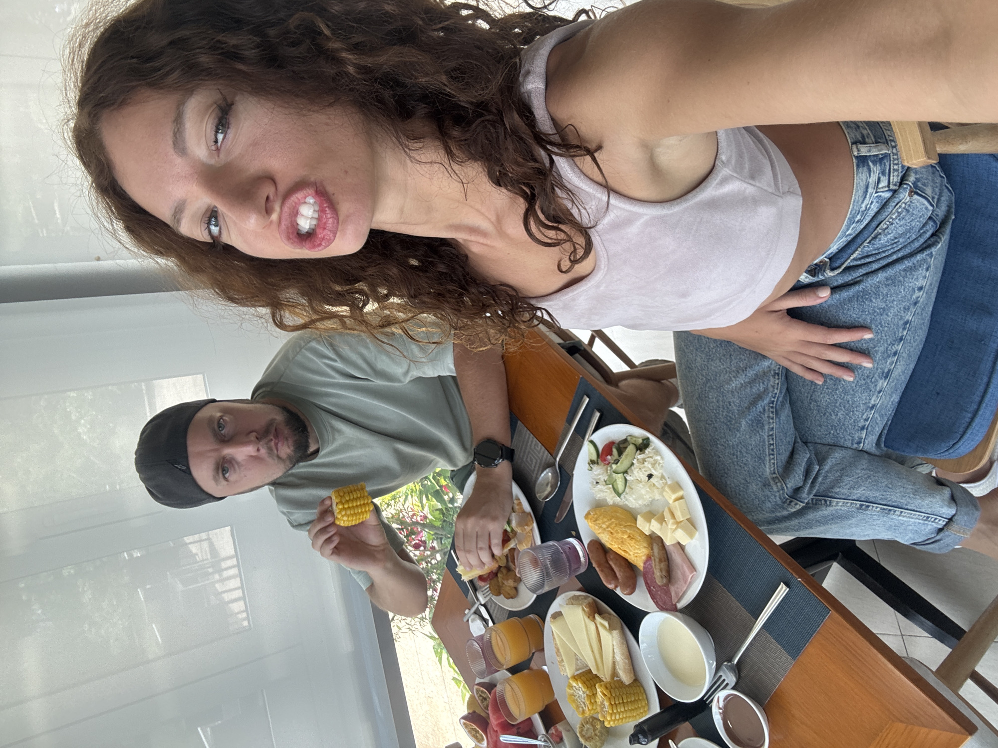 A photo shows a couple posing for a selfie at a breakfast table. The woman, fair-skinned with curly brown hair, is making a funny face. She wears a light pink crop top and blue jeans. Her right hand rests on her stomach. A man with short hair and a black beanie sits beside her. He holds a piece of corn on the cob.
The table is set with a variety of breakfast items including corn on the cob, cheese, sausages, watermelon, and juice. There are plates of salad and bread.
The background is bright, with a glimpse of greenery through a window. The location is likely Đà Nẵng, Vietnam. The photo appears to be taken indoors, possibly in a hotel or restaurant.