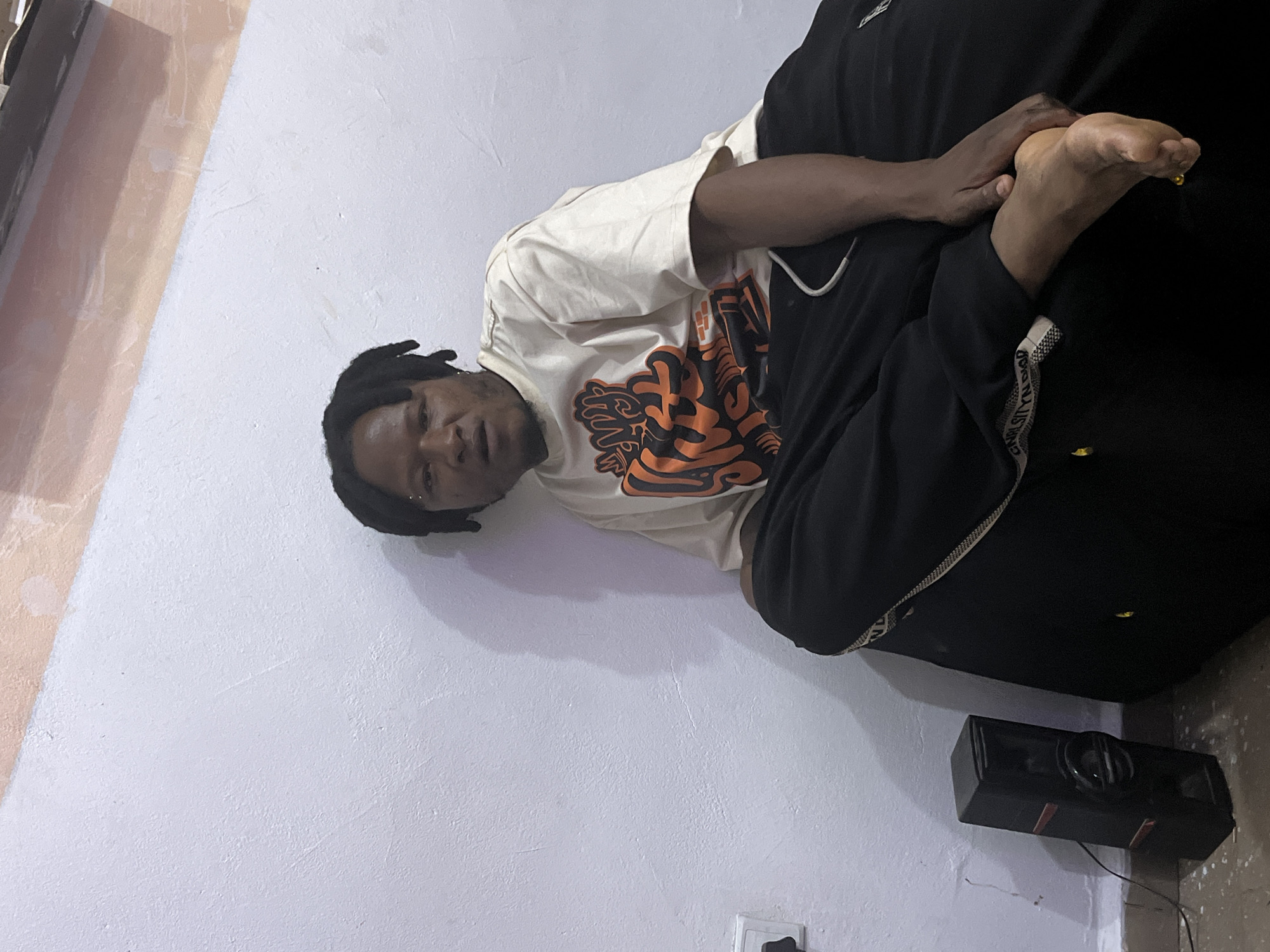 A person with dreadlocks is sitting on a black cushioned object next to a white wall. The person is wearing a light-colored t-shirt with an orange graphic and black pants. Their right foot is visible, and their hand is resting on it. To the left of the person, a black speaker is leaning against the wall. A white electrical outlet is also visible on the wall. The wall has some textured imperfections and a band of light orange and pink at the top. The floor appears to be concrete with some debris. The scene is indoors, likely in a residential or studio setting in Jos, Nigeria.
