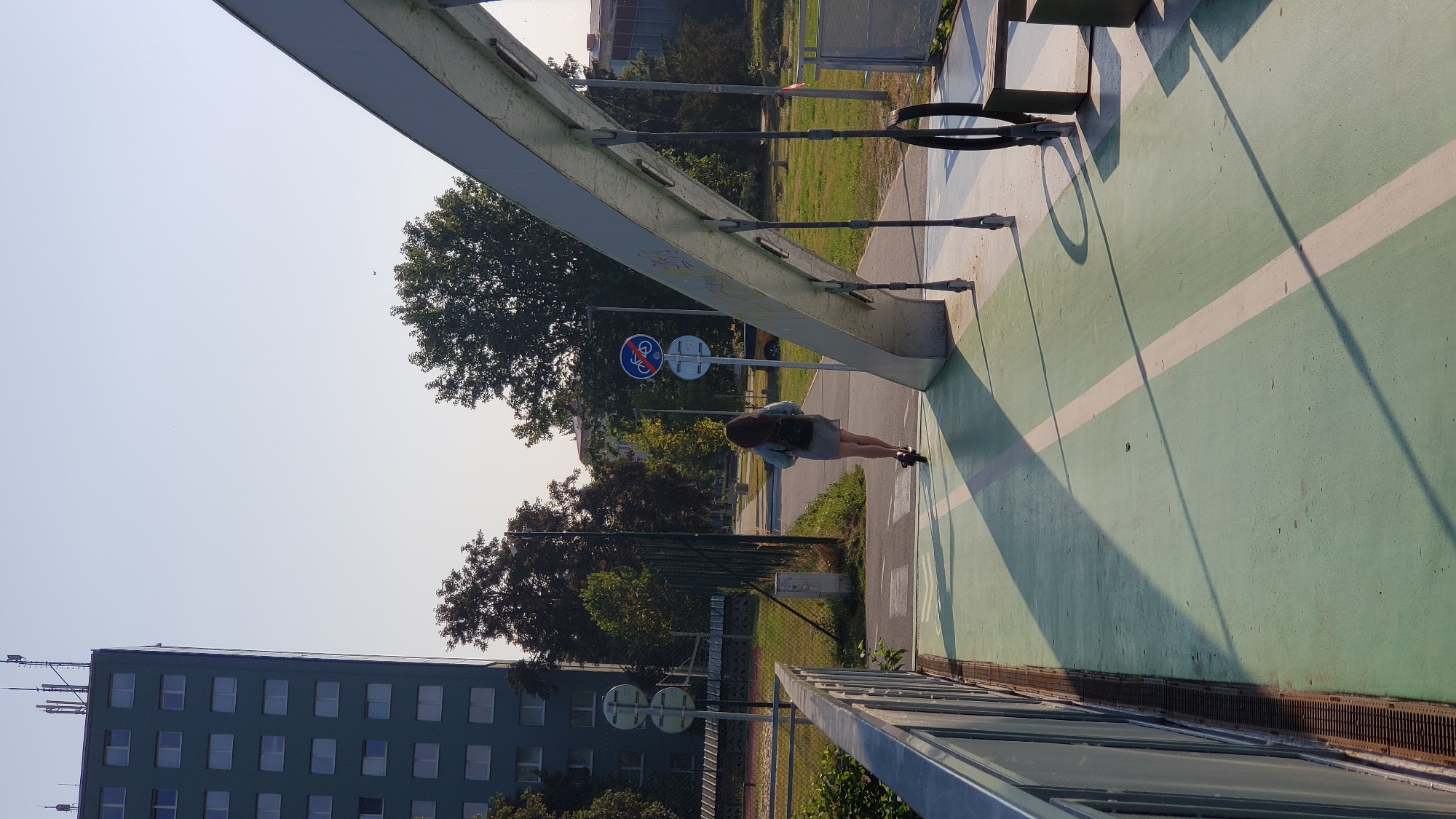 The image depicts a person riding a scooter on a green pedestrian and bicycle path, elevated by a bridge structure. The bridge has a grey metal railing on the left and a prominent grey metal archway supporting the structure on the right. The path is marked with white lines. In the background, a tall, dark green building with multiple windows stands to the left, and trees line the path. Traffic signs, including one indicating no cycling, are visible. The scene appears to be in an urban environment, likely Prešov, Slovakia, given the context. The time of day suggests it is either morning or late afternoon, due to the long shadows cast by the bridge structure and a solitary tree. A few other minor structures and grassy areas are also visible.