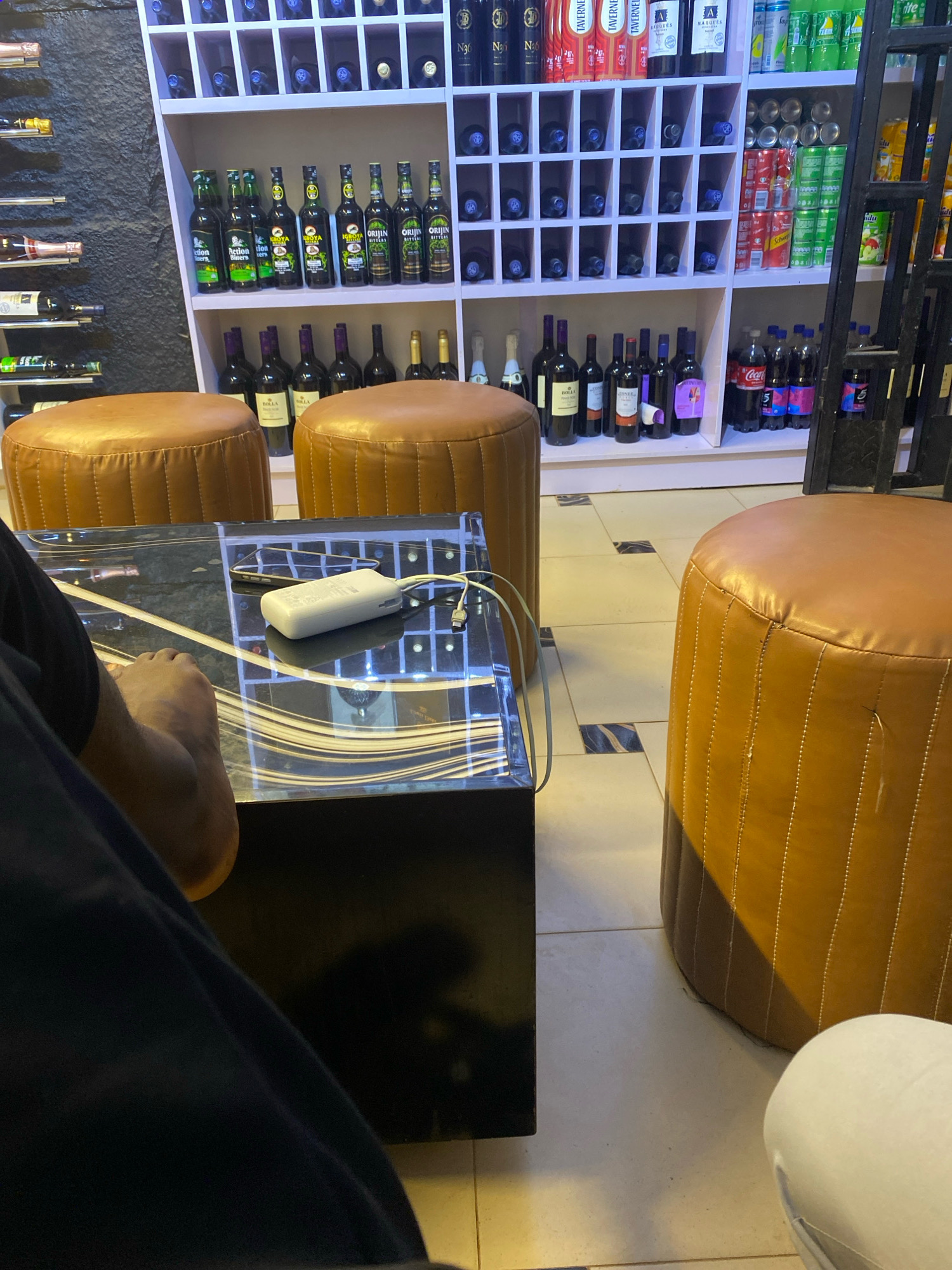 This is a forensic, matter-of-fact description of a scene that appears to be inside a liquor store in Jos, Nigeria. The image captures a portion of the store's interior, focusing on a display shelf stocked with various beverages and two plush, brown, round stools in the foreground.
The most prominent feature of the scene is the expansive shelving unit filled with an assortment of drinks. On the top shelf, there are rows of bottled wines and spirits, with some labels partially visible. Below this, a section is dedicated to individual wine bottles, neatly arranged in cubbies. Further down, there are bottles of what appear to be soft drinks or other beverages, including several cans of Coca-Cola and other brands. The shelves are illuminated by overhead lighting, casting a warm glow on the products.
In the foreground, two round, cushioned stools made of what looks like brown leather are positioned in front of the shelving unit. The stool on the left is partially obscured by a dark-colored fabric, likely clothing. On the surface of a glass-topped table between the stools, a white rectangular object, possibly a power bank or portable charger, is visible alongside a smartphone. A white cable extends from the power bank. The table's surface has an intricate, reflective design.
A person's leg and foot are visible in the lower left of the frame, indicating their presence in the scene, seated or resting. Their foot is bare. The flooring consists of light-colored tiles with dark, rectangular accents. The overall environment suggests a commercial establishment designed for the sale of alcoholic and non-alcoholic beverages. The presence of various brands of drinks, shelving units, and seating for customers are indicative of a well-stocked liquor store. The specific location within Jos, Nigeria, is inferred from the context of typical commercial establishments in that region.