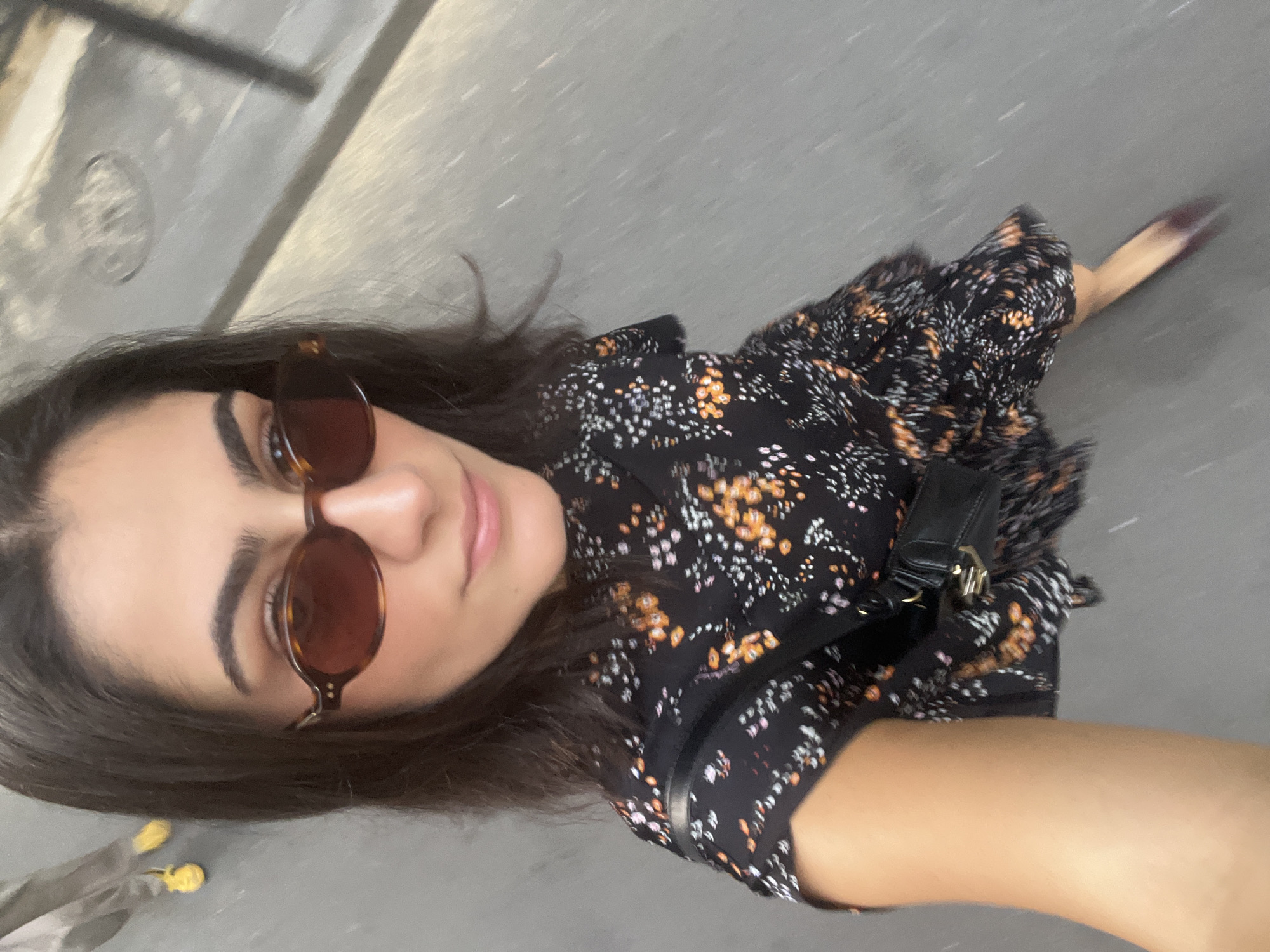 A woman in a black floral print dress and sunglasses is walking on a street in Paris, France. The street appears to be paved with grey asphalt and there is a drain cover visible in the background. The woman is wearing a small black crossbody bag. Her hair is dark brown and she has on light makeup. Another person's legs wearing yellow shoes are visible in the upper left corner of the image. The image is taken from a high angle, looking down at the woman. The lighting is natural and it appears to be daytime.
