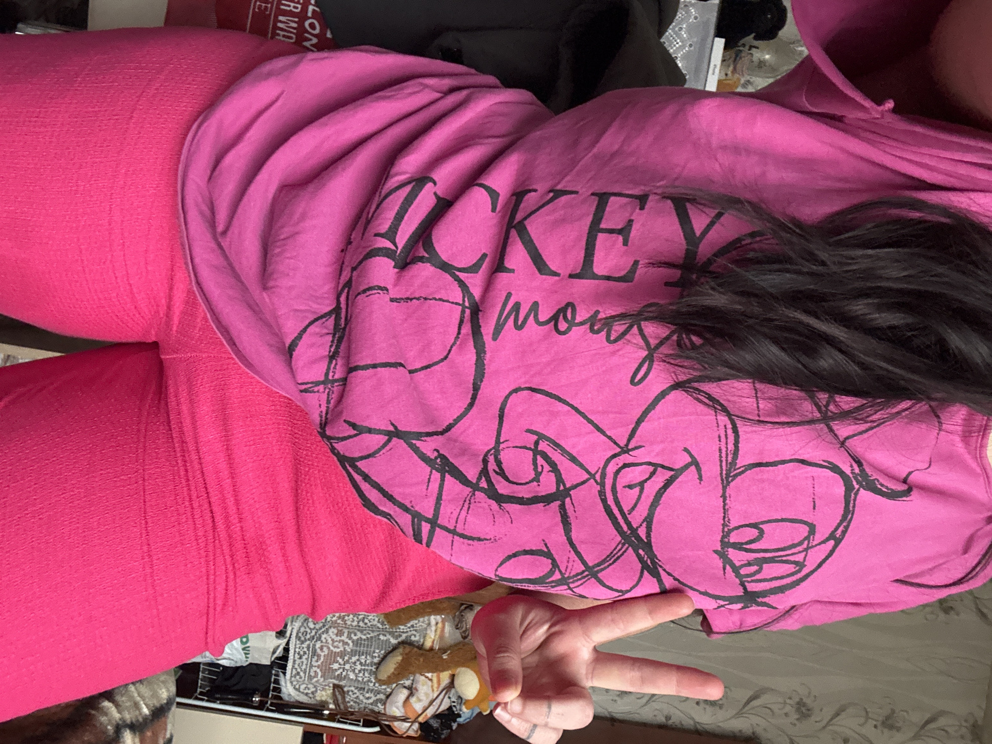 A person wearing pink leggings and a pink hooded shirt with a "Mickey Mouse" design is holding a peace sign. The background includes household items, suggesting an indoor setting. The location is not discernible as Dobrovnitsa, Bulgaria from the image alone.