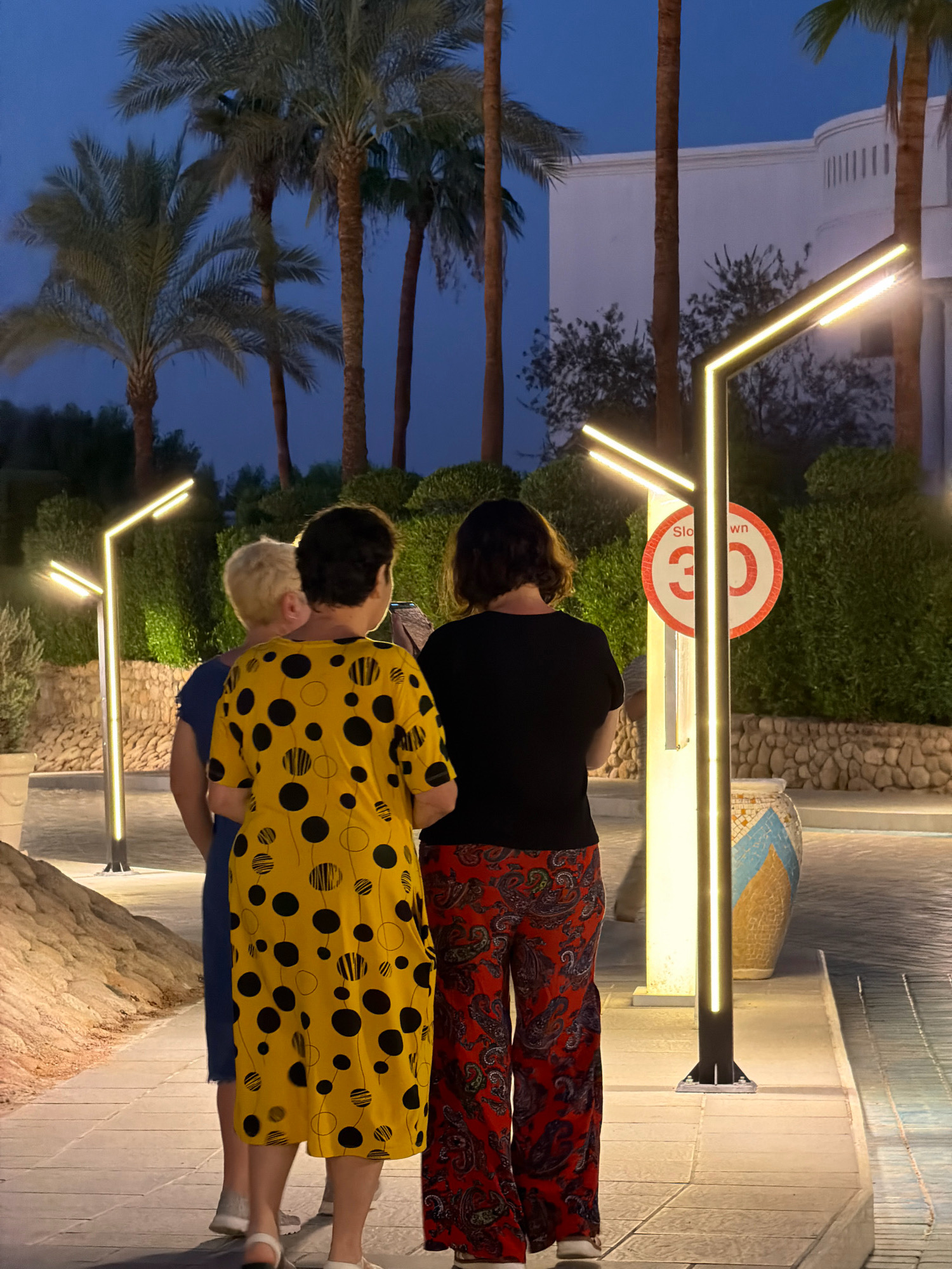 Three individuals are standing on a paved walkway at dusk, facing away from the camera. The walkway is illuminated by modern, vertical light fixtures that emit a warm, yellow glow. Palm trees and lush green shrubbery are visible in the background, with a white, multi-story building in the distance. A circular speed limit sign, indicating 30, is attached to one of the light poles. The ground is paved with light-colored tiles, and a decorative, mosaic-patterned pot sits to the right of the walkway. The scene appears to be in a resort or hotel area, possibly in Sharm El Sheikh, Egypt, given the palm trees and architecture.