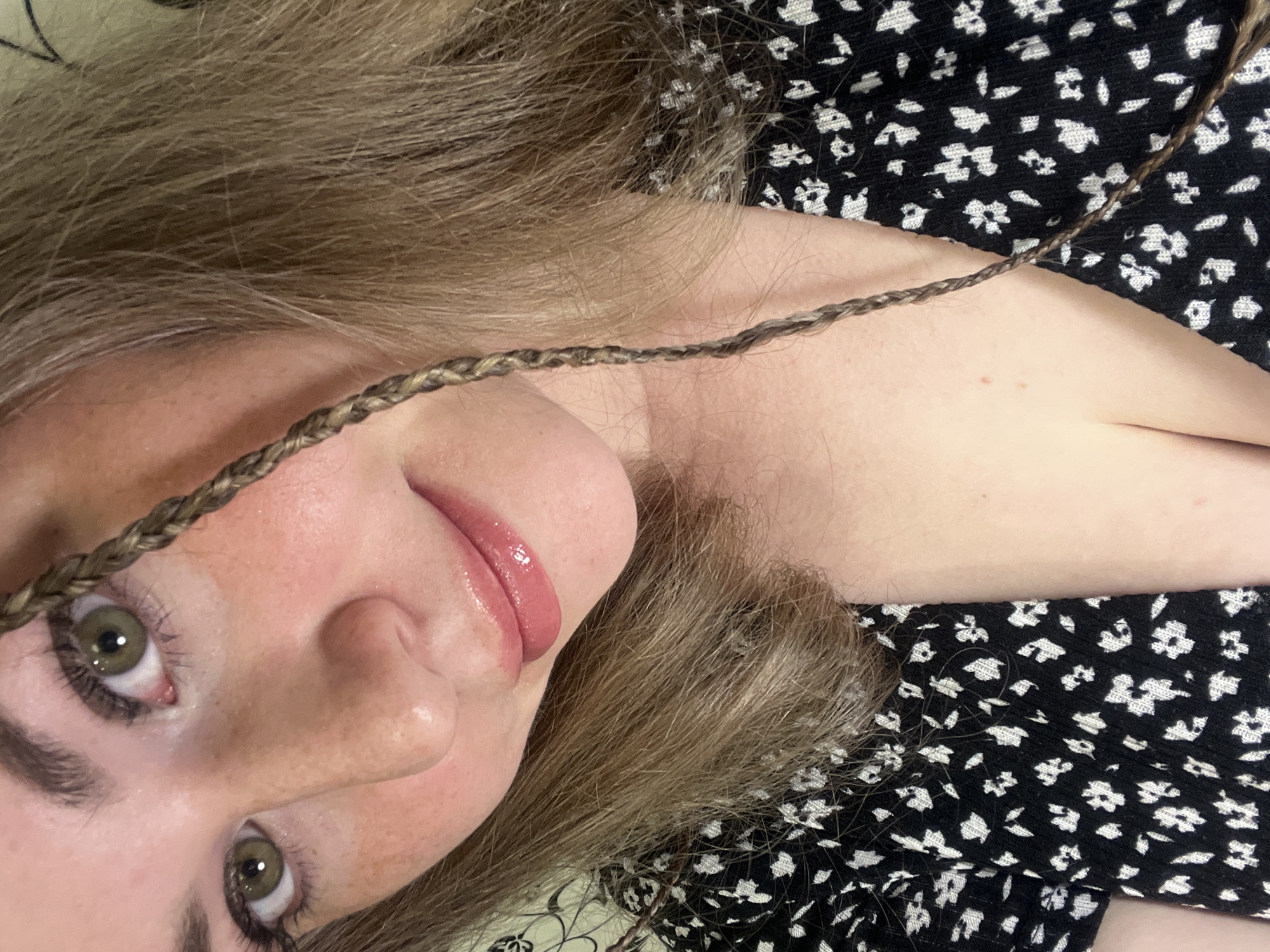 A close-up, slightly angled shot shows a person with fair skin and light brown hair lying down. A thin braid of hair extends across the frame, near the person's face. The person's eyes are green and appear to be looking at the camera. The lips have a glossy pink color. The person is wearing a black and white floral-patterned top. No identifiable location in Chaikovsky, Russia can be determined from the image. The environment is out of focus and appears to be a light-colored surface.