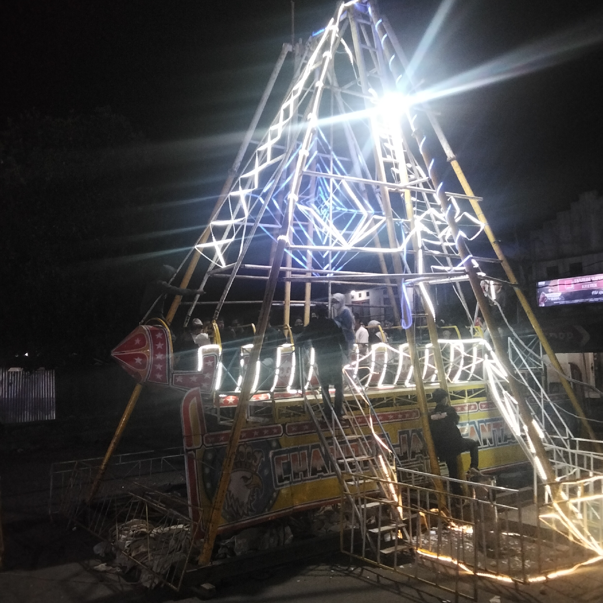 The image shows a brightly lit amusement ride at night in Cisaat, Indonesia. The ride, which appears to be a swing ride with a large central structure, is adorned with numerous white and blue lights, creating a dazzling display against the dark sky. Several people are visible within the ride's cabins, and others are standing on or near the structure, possibly operating or observing the ride. The ground around the ride is cluttered with debris, including metal scaffolding and discarded materials, suggesting an outdoor event or a temporary setup. In the background, a building with a billboard featuring a cigarette advertisement can be seen. The overall scene depicts a lively atmosphere at a local fair or amusement park.