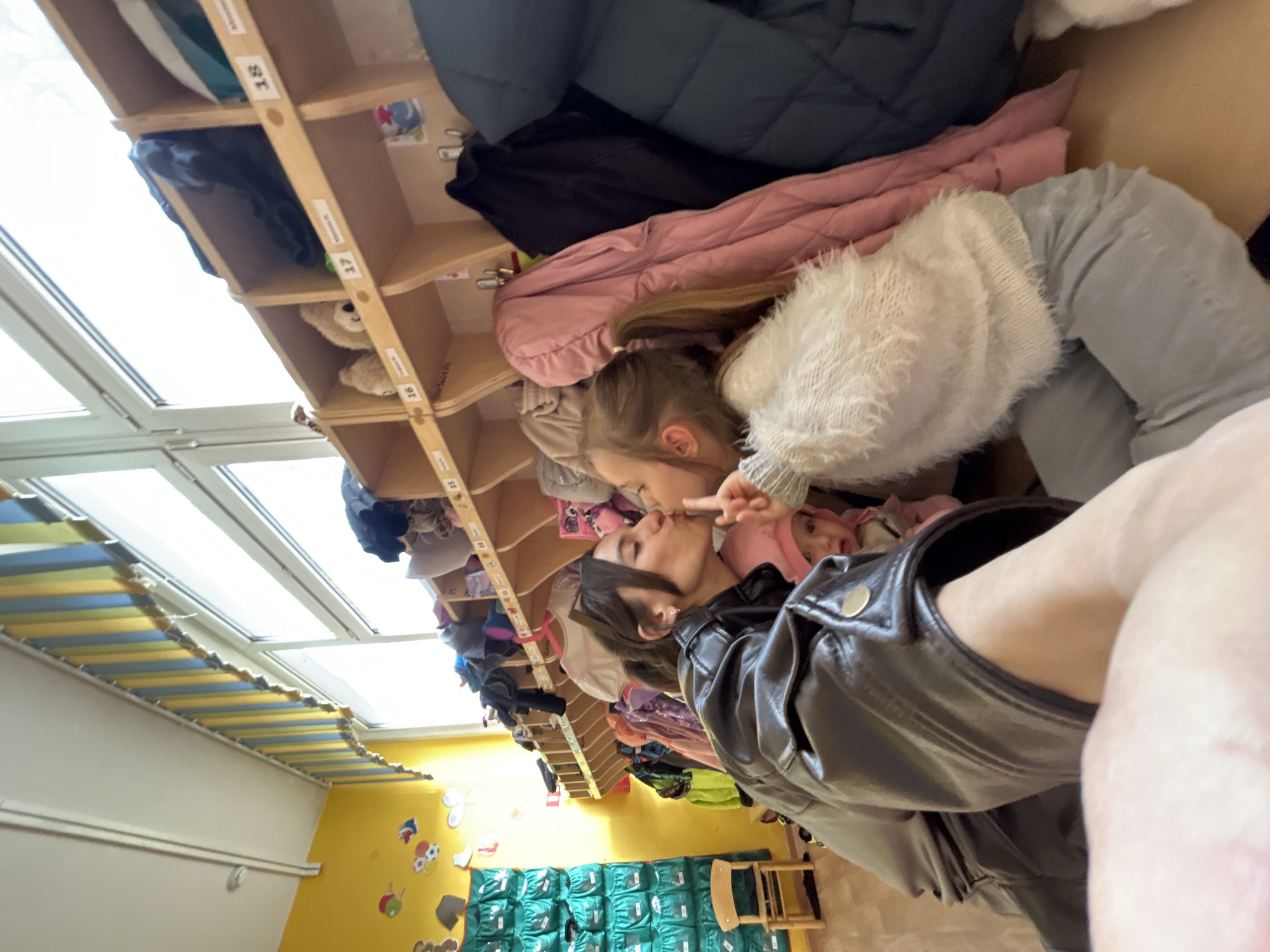 The image shows a scene in a children's daycare, likely in Moscow, Russia, given the presence of children's lockers with Cyrillic names on them. A girl with blonde hair in a ponytail, wearing a white sweater and gray pants, is reaching out to touch the face of a woman wearing a black leather jacket. The woman is leaning down, her face close to the girl's. The background shows rows of lockers, some containing children's clothing like coats and jackets. The room has a yellow wall with colorful decorations and windows in the ceiling. There is also a shelf unit filled with green bags and a child's chair. A baby in a pink outfit is visible next to the woman.