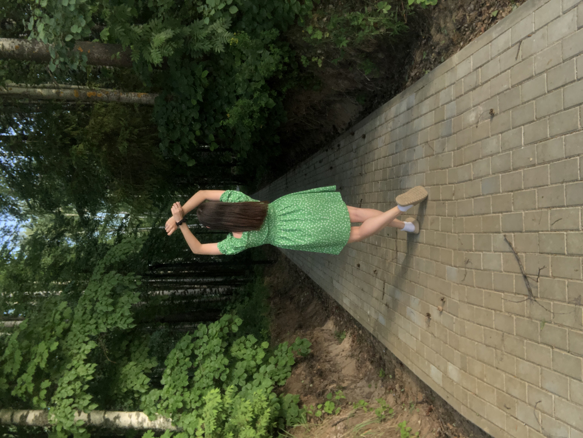 A woman in a short, green, floral dress is seen from above, lying on a brick-paved pathway in a wooded area. The woman has long, dark hair and is wearing light-colored shoes. Her arms are raised above her head. The pathway is bordered on both sides by lush green trees and shrubs. The location is Noviy, Russia.