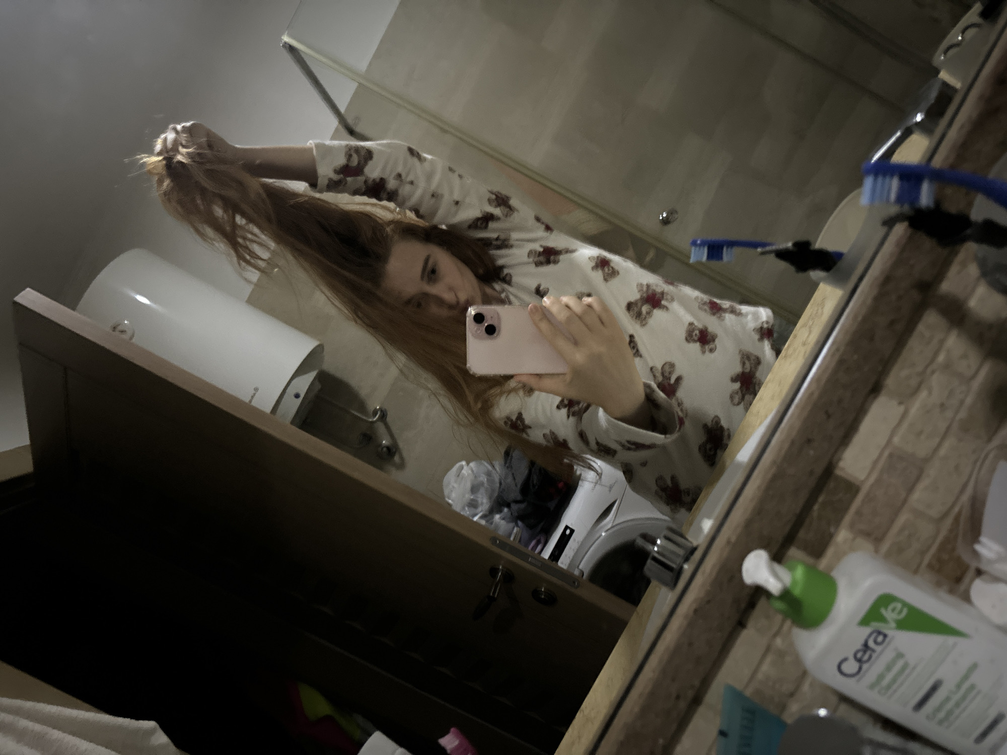 A woman is taking a selfie in a bathroom, likely in an apartment or hotel in Budva, Montenegro. She is holding up a large section of her long, reddish-brown hair with one hand, while holding a pink smartphone in the other hand to take the picture. She is wearing pajamas with a teddy bear pattern.
The bathroom has a sink with a Cerave moisturizing cream bottle on the countertop. A toothbrush and other toiletries are also visible. Below the sink is a cabinet with an open drawer. In the background, a washing machine is visible. The walls appear to be tiled, and a showerhead is visible on the wall. A white cylindrical object, possibly a water heater, is mounted above the cabinet.