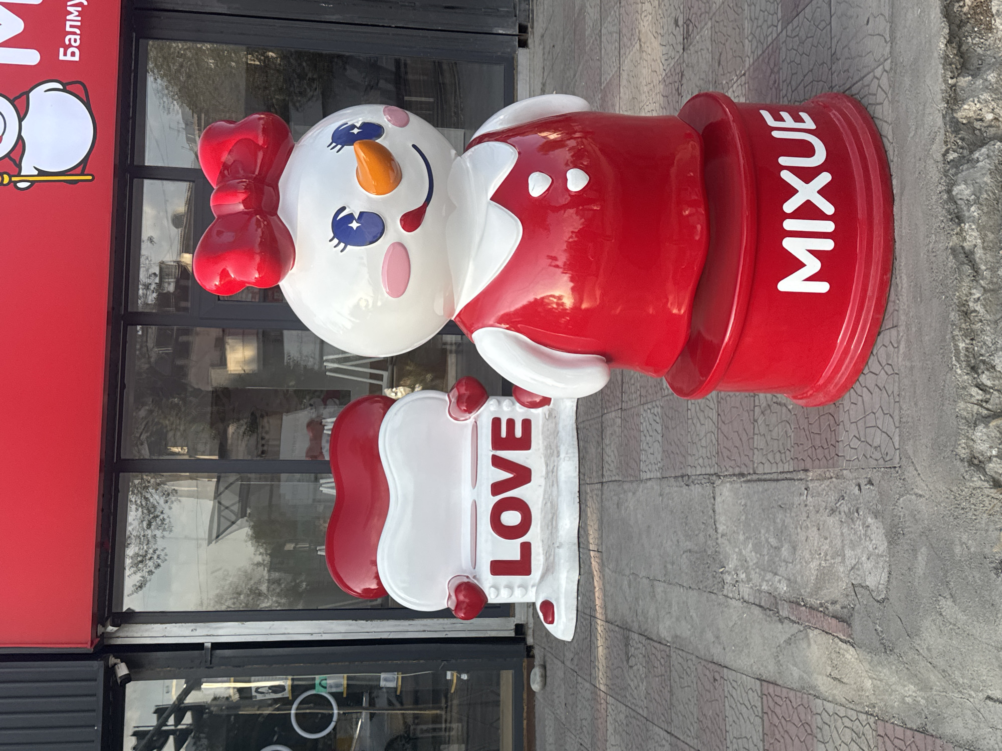 In Bishkek, Kyrgyzstan, a red and white MIXUE storefront features a whimsical outdoor display. A large, stylized snowman figure, adorned with a bright red bow and a red dress, stands prominently. Beside it, a white bench shaped like stacked red hearts with the word "LOVE" in red letters invites passersby. The figures are positioned on a paved sidewalk in front of the store, with reflections visible in the large glass windows behind them. The scene is captured during daylight hours.