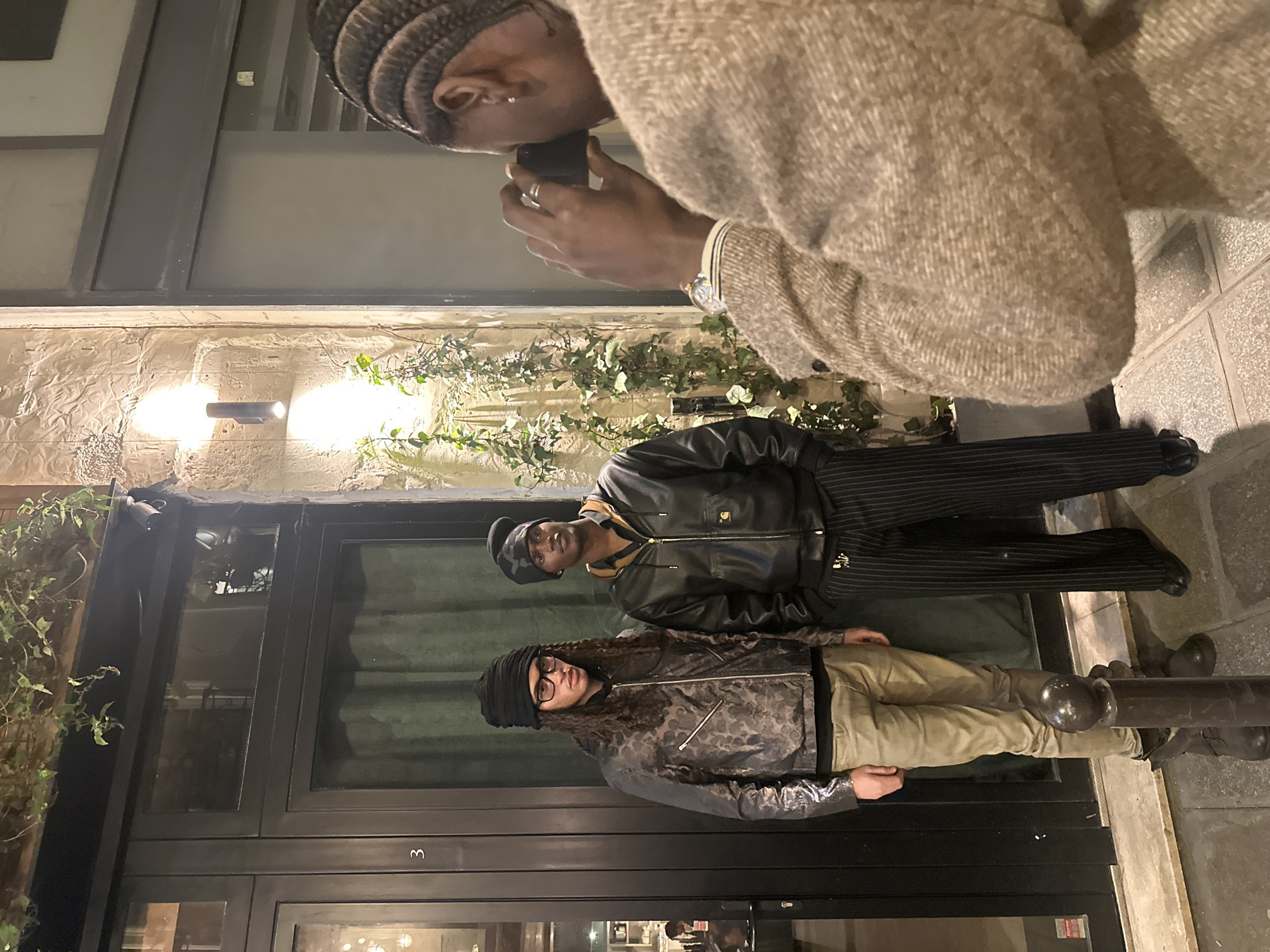 An urban street scene in Paris, France, at night depicts three males. In the foreground, partially visible on the right, a male with braided hair and a beige textured coat holds a black smartphone horizontally, aiming it to photograph the two males positioned in the mid-ground. He wears a watch on his left wrist and a silver ring on his left ring finger.
The two subjects of the photograph stand against a dark-framed storefront with green curtains visible through the glass. The left individual, a male with long, dark curly hair and clear-framed eyeglasses, wears a black knitted beanie, a dark patterned bomber jacket, a black undershirt, khaki pants, and dark boots. He stands with his hands at his sides. The right individual, a male of Black/African descent, wears a dark knitted beanie, a black leather-like bomber jacket with a brown collared shirt beneath, dark pinstriped trousers, and dark shoes. He stands with his hands in his pockets.
The building facade is composed of light-colored, textured stone, illuminated by two upward and downward-facing sconce lights. Green ivy grows on the stone wall and above the storefront. The storefront features a dark frame with a glass door displaying the number "3" and a window above it. A small white security camera is mounted above the right side of the storefront window. A dark, spherical-topped bollard is present on the paved sidewalk in front of the subjects. The ground is a tiled pavement.