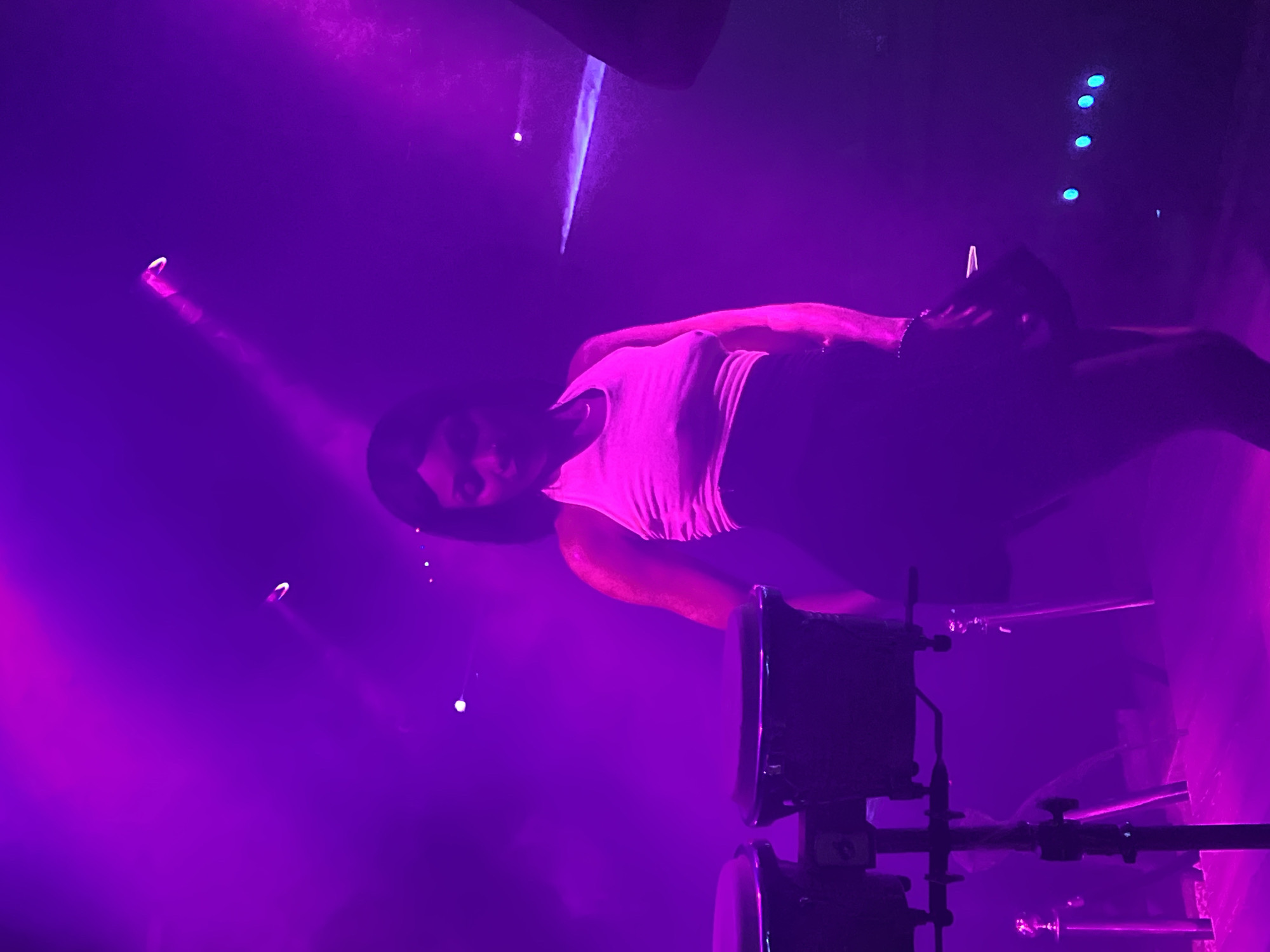 A woman in a white halter top and black skirt is performing on a stage in a club in Howayi, Nigeria. She is wearing a necklace and bracelets and her hair is black and short. The stage is lit with purple and pink lights, and there is a drum set in the background. The woman is dancing and her body is angled to the left. The lighting creates a hazy effect, and there are a few small blue lights on the right side of the stage. The scene is captured at eye level.