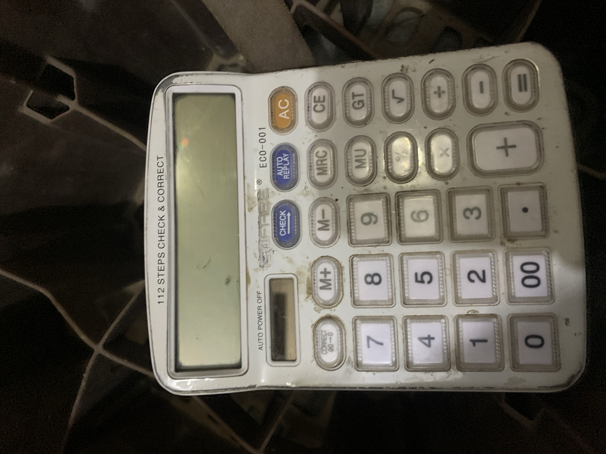 The image shows a close-up, top-down view of a dusty, white calculator situated on a dark, textured surface, possibly a woven material or the interior of a bag. The calculator is a standard model with a digital display at the top, which is blank and appears smudged. Below the display are various buttons for numerical input (0-9, 00), arithmetic operations (+, -, x, ÷, =), memory functions (M+, M-, MRC), and other functions like CHECK, AUTO REPLAY, AC, CE, GT, MU, %, and √. The calculator has markings indicating "112 STEPS CHECK & CORRECT" and "AUTO POWER OFF". The object appears to be in a used condition, with visible dust and grime on its surface and the surrounding area. The environment is dark and out of focus, providing little detail about its surroundings. There is no discernible information about the location being New Stadium, Nigeria, or any people present in the image.