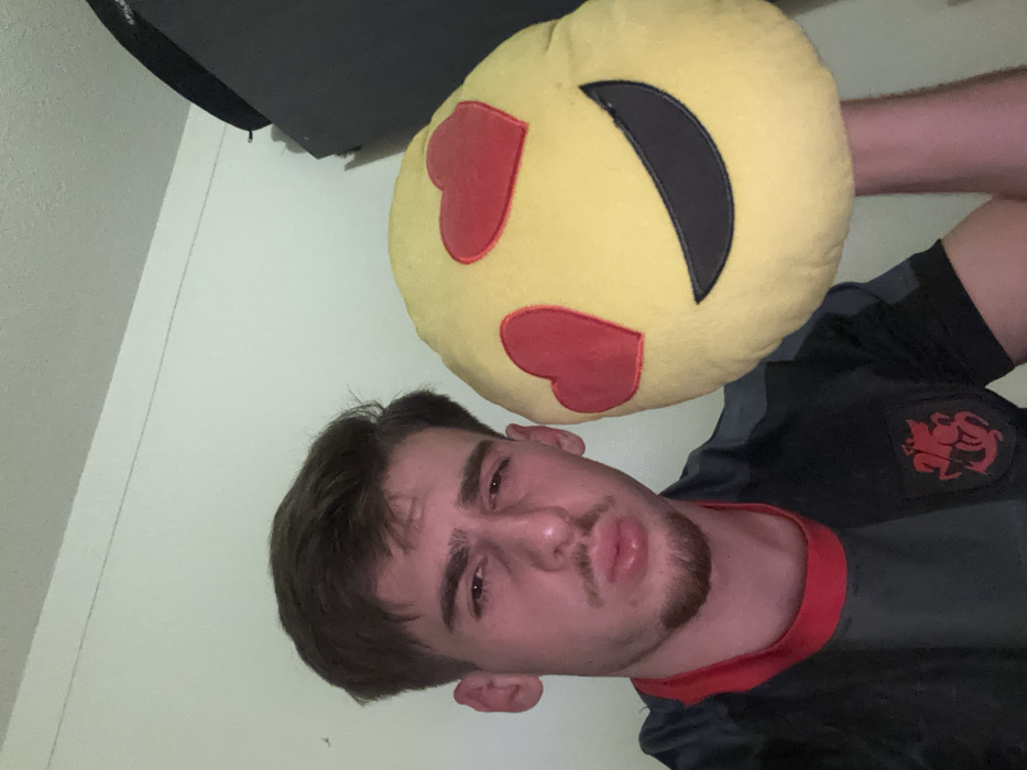 A young man with short brown hair, light skin, and a goatee is positioned in the lower half of the frame, looking at the camera with a slightly furrowed brow and pursed lips. He's wearing a gray and black athletic shirt with red trim. Above him, partially obscuring the top right of the frame, is a yellow emoji pillow featuring heart eyes and a smirk. The background is a plain, light-colored wall with a dark object in the top left corner. The image is taken from a slightly low angle.