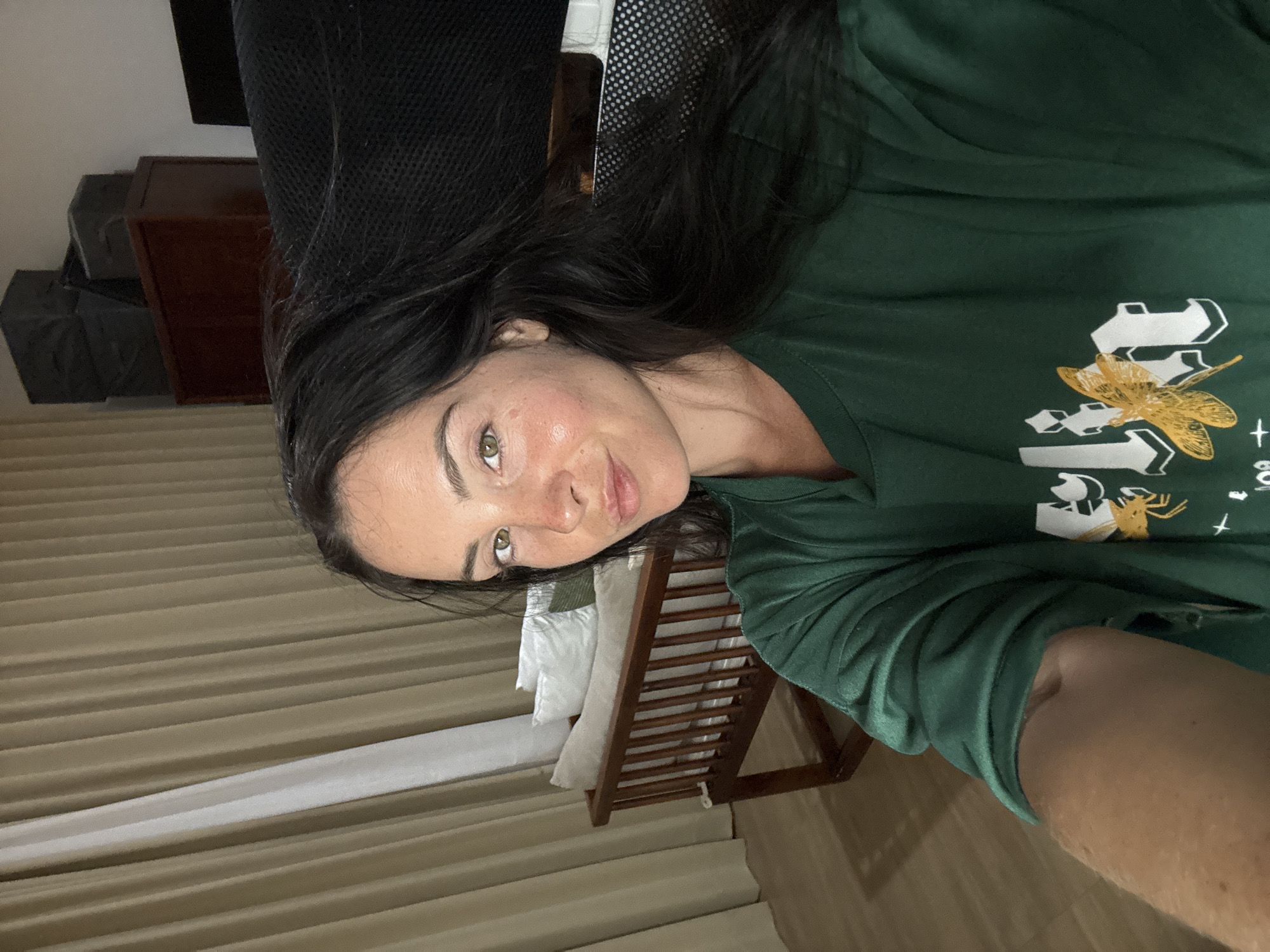 The image shows a close-up, selfie-style shot of a woman with long, dark hair and light green eyes. She is wearing a dark green t-shirt with a white graphic featuring a yellow dragonfly. The woman has light skin with some freckles and a slightly flushed complexion. She is looking directly at the camera with a subtle smile.
The background appears to be an indoor setting, likely a bedroom or living area. Behind the woman, there are floor-to-ceiling beige curtains partially drawn, revealing a white sheer curtain on the left. To the right of the curtains, there is a wooden bench with white cushions and a wooden piece of furniture with drawers. Stacked on top of this furniture are several dark grey boxes. Further to the right, a dark, textured surface, possibly a chair or sofa, is visible. The flooring is light-colored wood.
While the image itself does not explicitly indicate the location of North Kuta, Indonesia, this description pertains to the visual elements present in the photograph.
