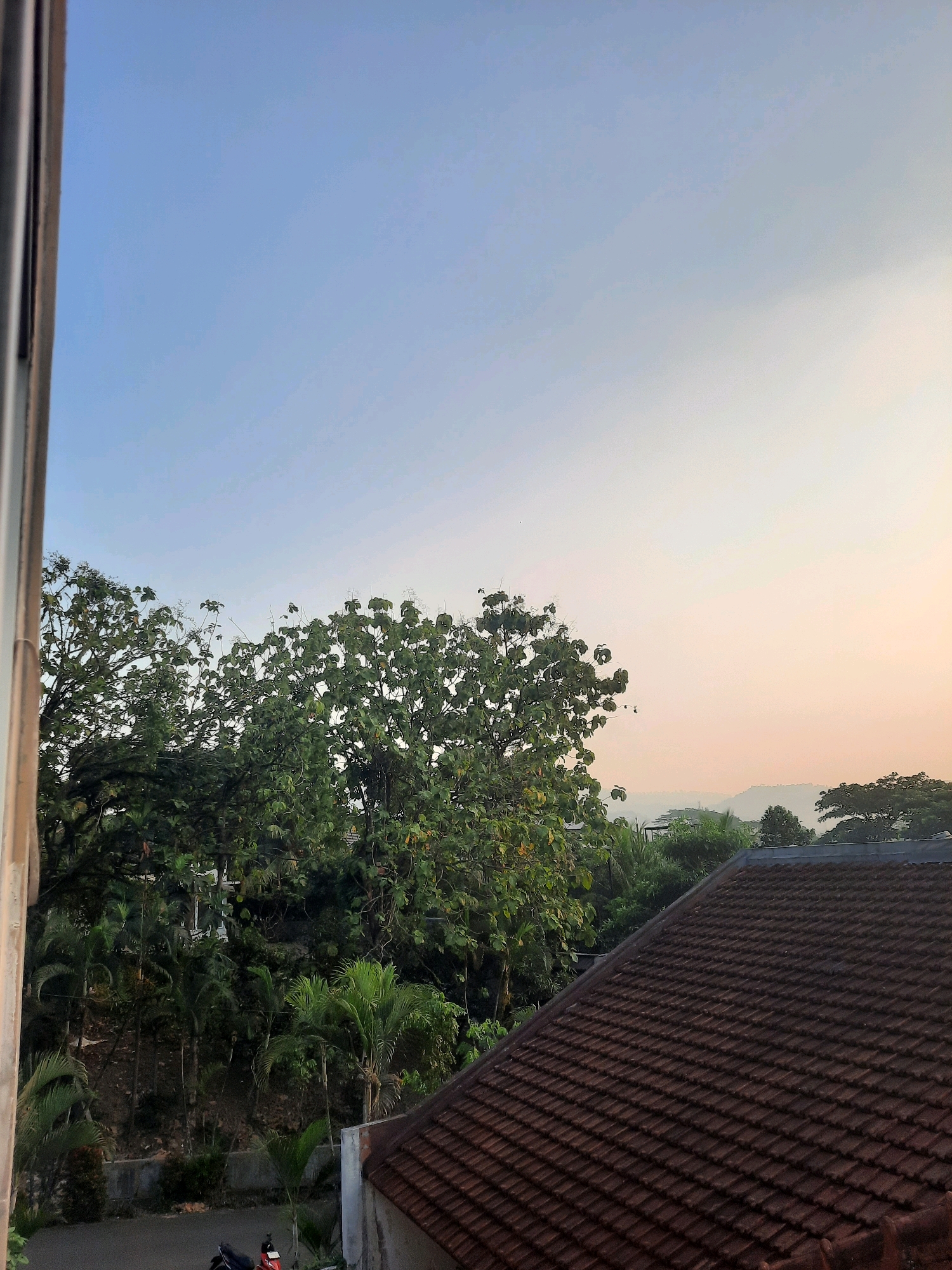An exterior, medium shot captures a dawn or dusk scene viewed from a window or elevated vantage point in Babakan Madang, Indonesia. The sky transitions from a clear light blue at the top to a soft orange-pink hue near the horizon. Lush green foliage, primarily large trees with broad leaves and smaller palm trees, dominates the middle ground. In the foreground, a tiled roof with weathered, reddish-brown tiles angles diagonally across the lower right portion of the frame. The left edge of the image shows the side of a building with a light-colored wall. A dark asphalt road is visible at the bottom left, with a red and black motorcycle parked on it. The overall atmosphere suggests a tranquil, natural environment within a semi-urban or suburban setting.