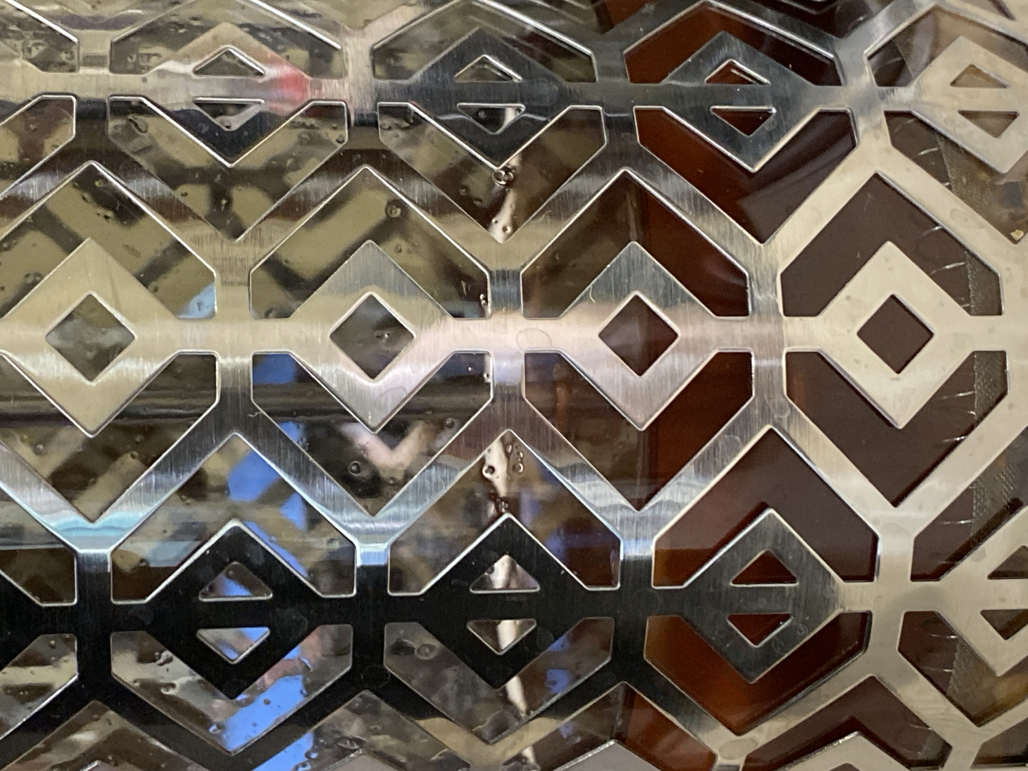The image shows a close-up, top-down view of a metallic decorative grate with a geometric pattern. The grate is silver and appears to be made of brushed metal, exhibiting reflections of its surroundings. The pattern consists of interlocking V-shapes and diamond shapes, creating a repeating tessellation. The background, visible through the openings of the grate, is dark brown and appears to be a solid surface.
The metallic surface of the grate has some minor imperfections, including small circular marks and what appear to be water droplets or smudges. These imperfections, along with the reflections, suggest the grate is part of an object or structure in an environment with light sources and possibly liquid present. The reflections themselves are varied, showing patches of blue (possibly sky or a blue object) and lighter, diffused areas.
The overall impression is of a textured and reflective surface with a repeating, angular design. There is no discernible activity or event taking place; the image is a still life focusing on the texture and pattern of the grate. No text is visible in the image. The lighting appears to be ambient, with distinct reflections indicating multiple light sources or a complex environment. The lack of any specific environmental cues makes it impossible to determine the time of day, weather, or a precise location beyond the immediate context of the grate itself.