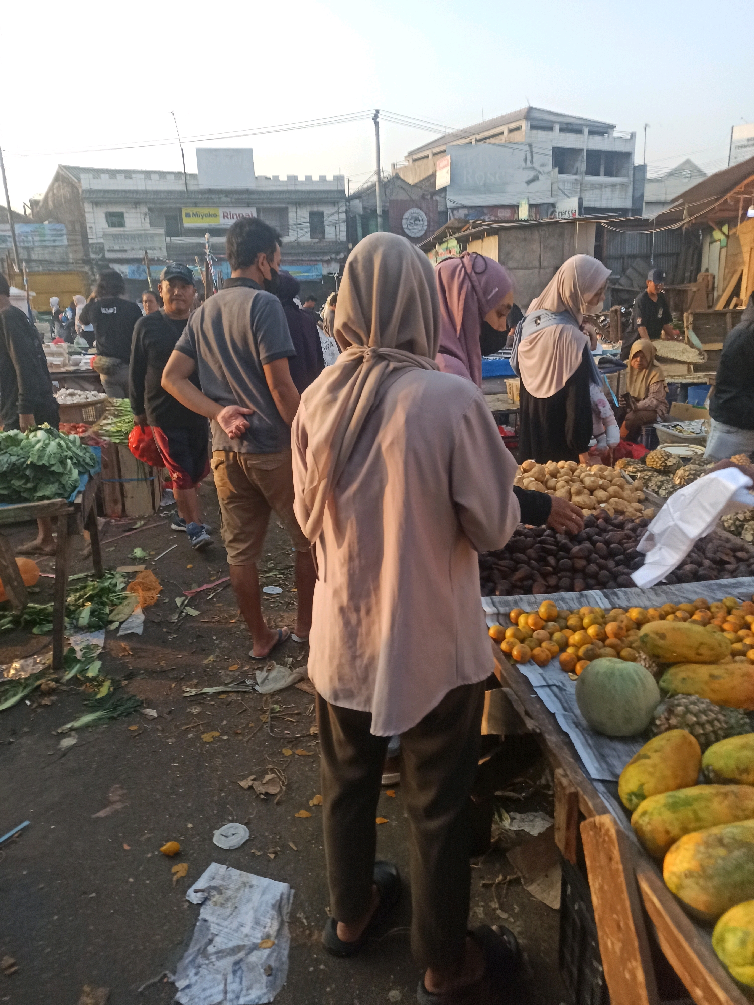 The image depicts an outdoor market scene in Cikarang, Indonesia, with a variety of fruits and vegetables on display. Several people are present, some wearing headscarves and masks, suggesting a public gathering. The environment appears to be a busy marketplace with stalls set up along a dirt path. In the foreground, a person in a light brown headscarf and beige shirt is seen from behind, facing a stall laden with various fruits, including tangerines, papayas, and pineapples. To the left, a stall with leafy green vegetables is visible. In the background, there are buildings and other market stalls, indicating a bustling commercial area. The lighting suggests it is either early morning or late afternoon.