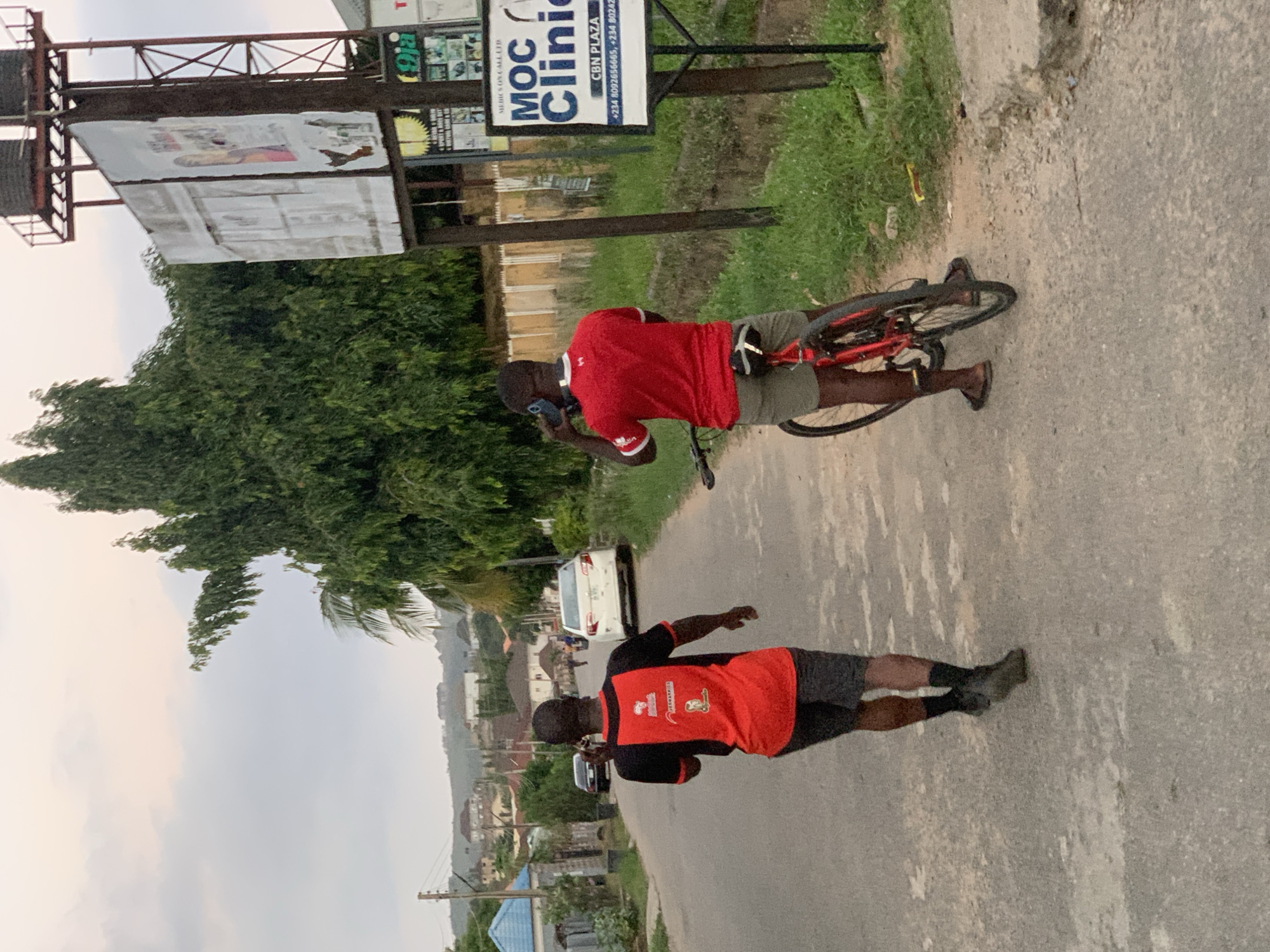 The image shows a street scene in Abuja, Nigeria. Two men are visible; one stands next to a red bicycle, using a mobile phone, wearing a red shirt with white trim and grey shorts. The second man is further down the road, also wearing a red shirt with a black yoke and grey shorts, with a logo or design on the back, and he may also be using a mobile phone. The MOC Clinic is visible on a signpost. An overhanging billboard and dense green trees are also present. There is a road with visible wear and tear, and a few buildings can be seen in the background. There's a white car parked on the side of the road.