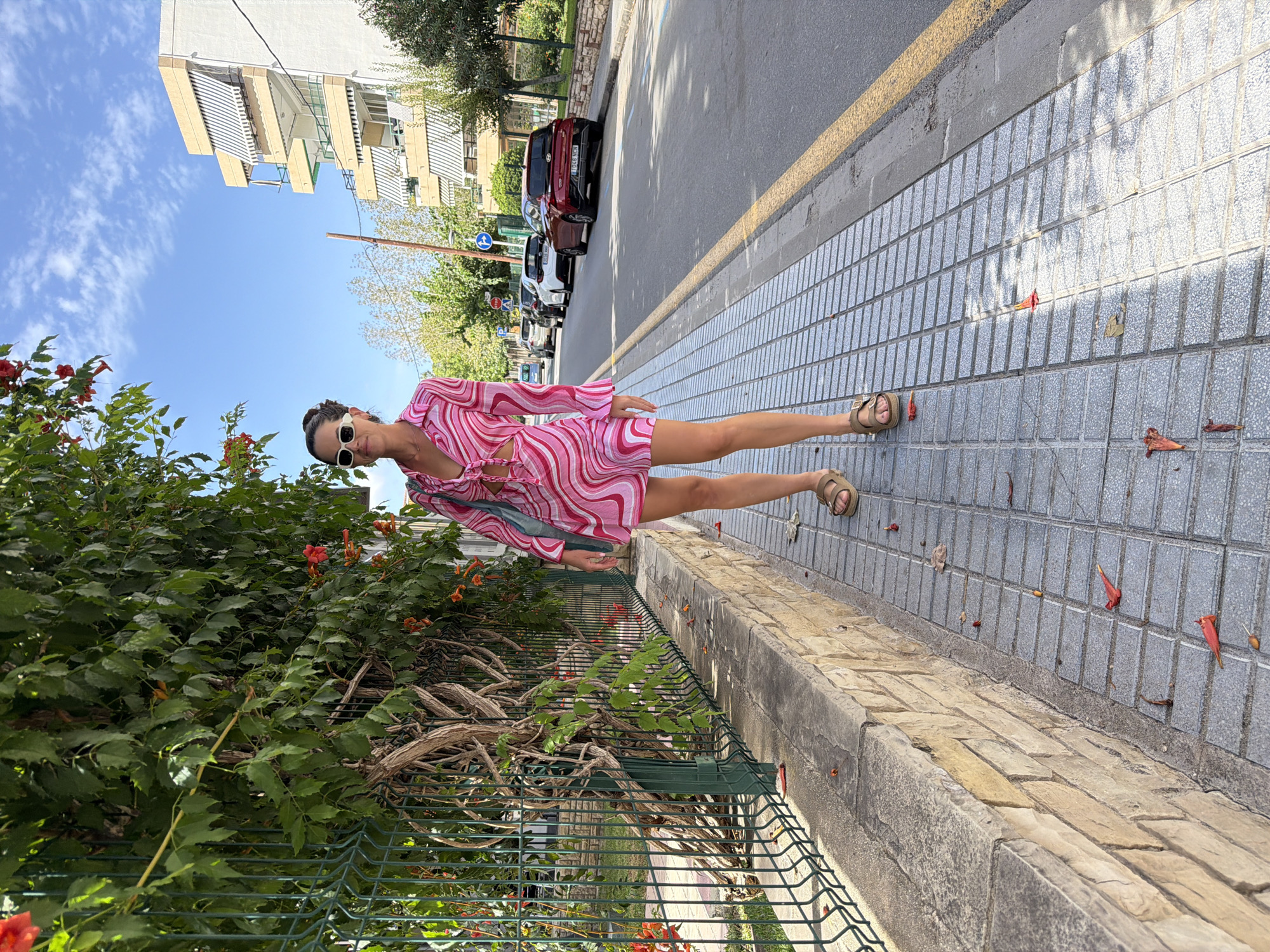 In Salou, Spain, on a sunny day, a woman with dark hair pulled back and wearing white sunglasses stands on a paved sidewalk. She is dressed in a pink and white swirl-patterned dress with cutouts at the waist, a tie-front neckline, and bell sleeves. She is also wearing brown Birkenstock-style sandals and carrying a teal bag over her shoulder. The woman is positioned on the sidewalk next to a low stone wall and a green metal fence, behind which is a wall of green foliage with some orange flowers. The street beside the sidewalk is lined with parked cars, including a red Hyundai and a white car. In the background, there is a multi-story building with balconies and striped awnings, along with trees and a clear blue sky with scattered clouds. The sidewalk has fallen orange flower petals scattered across it.