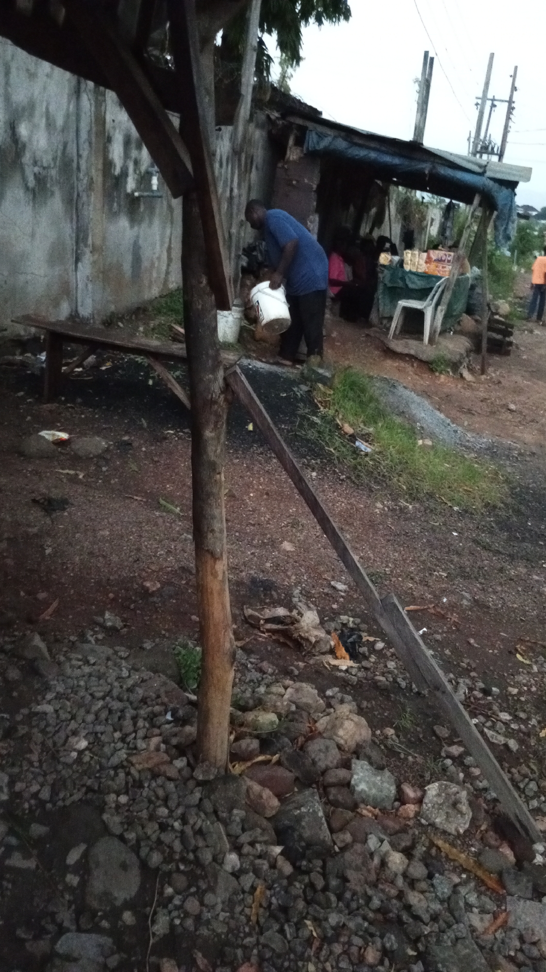 The scene depicts a makeshift structure in Adankolo, Nigeria. A man in a blue shirt is carrying a white bucket near a concrete building. The structure has a bench and appears to be a small shop with items on display. The ground is dirt and gravel with wooden support beams. Another person is visible standing in the distance. There are overhead electrical lines in the background.