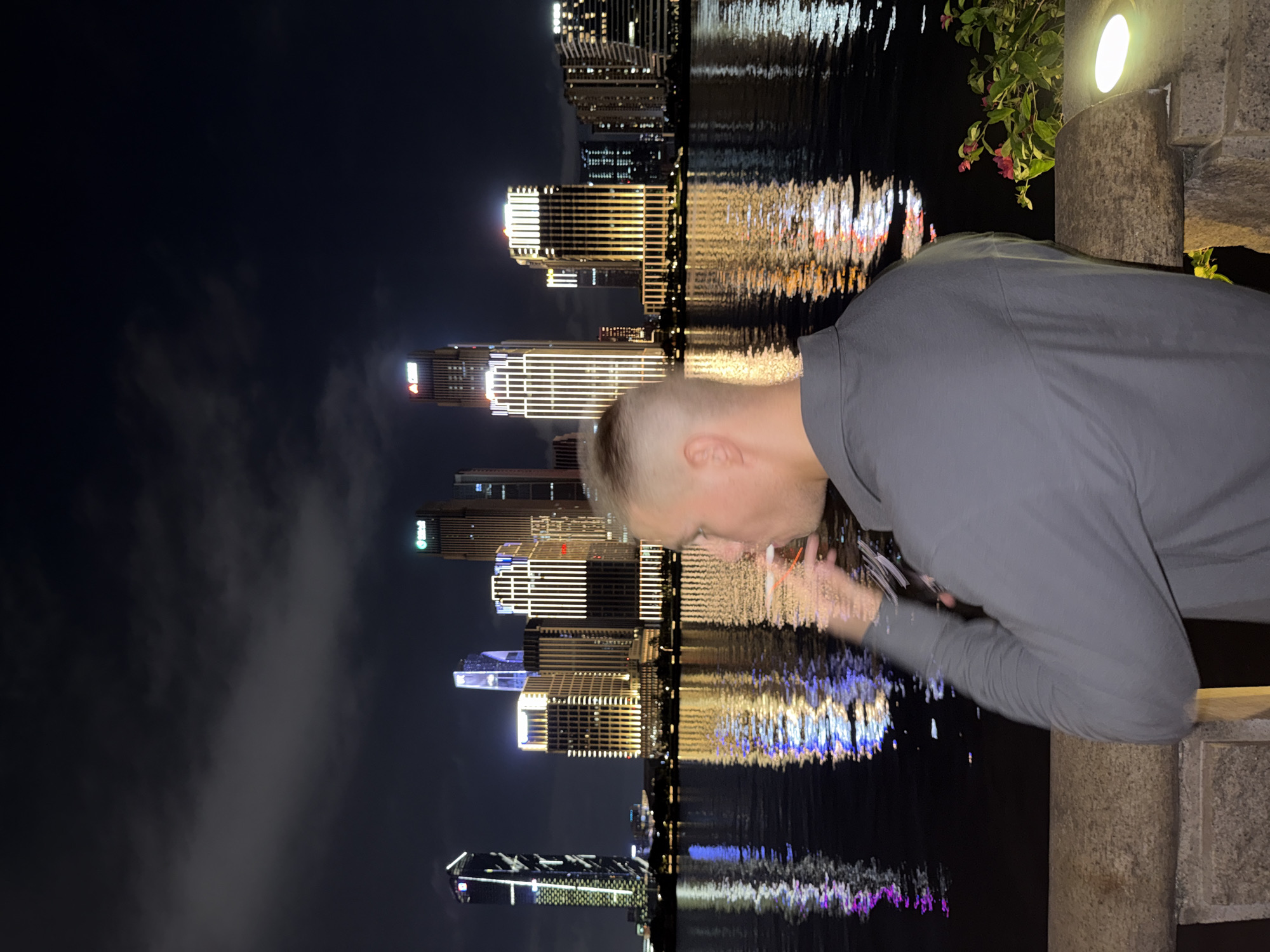 The image displays a nighttime urban waterfront scene in Tianhe District, China. In the foreground, a person is visible from the back and right side, wearing a grey long-sleeved collared shirt and possessing short hair. Their right hand is raised, holding a thin object to their mouth. A stone railing forms the immediate lower boundary of the foreground. To the right of the person, there is a cluster of green foliage with pink flowers and a ground-level light fixture emitting a warm yellow glow.
The middle ground is occupied by a dark body of water, reflecting the intense artificial lights from the distant city skyline as shimmering vertical streaks of white, gold, blue, and purple. The background is dominated by a dense array of modern skyscrapers and high-rise buildings, all extensively illuminated. Notable features include structures with prominent vertical gold-colored lighting schemes, a tall building on the far left exhibiting a distinct white and blue light pattern, and several buildings displaying corporate logos at their upper levels, including a green logo on a building within the right cluster and a red logo on another prominent skyscraper. The sky above the cityscape is dark with discernible cloud formations.